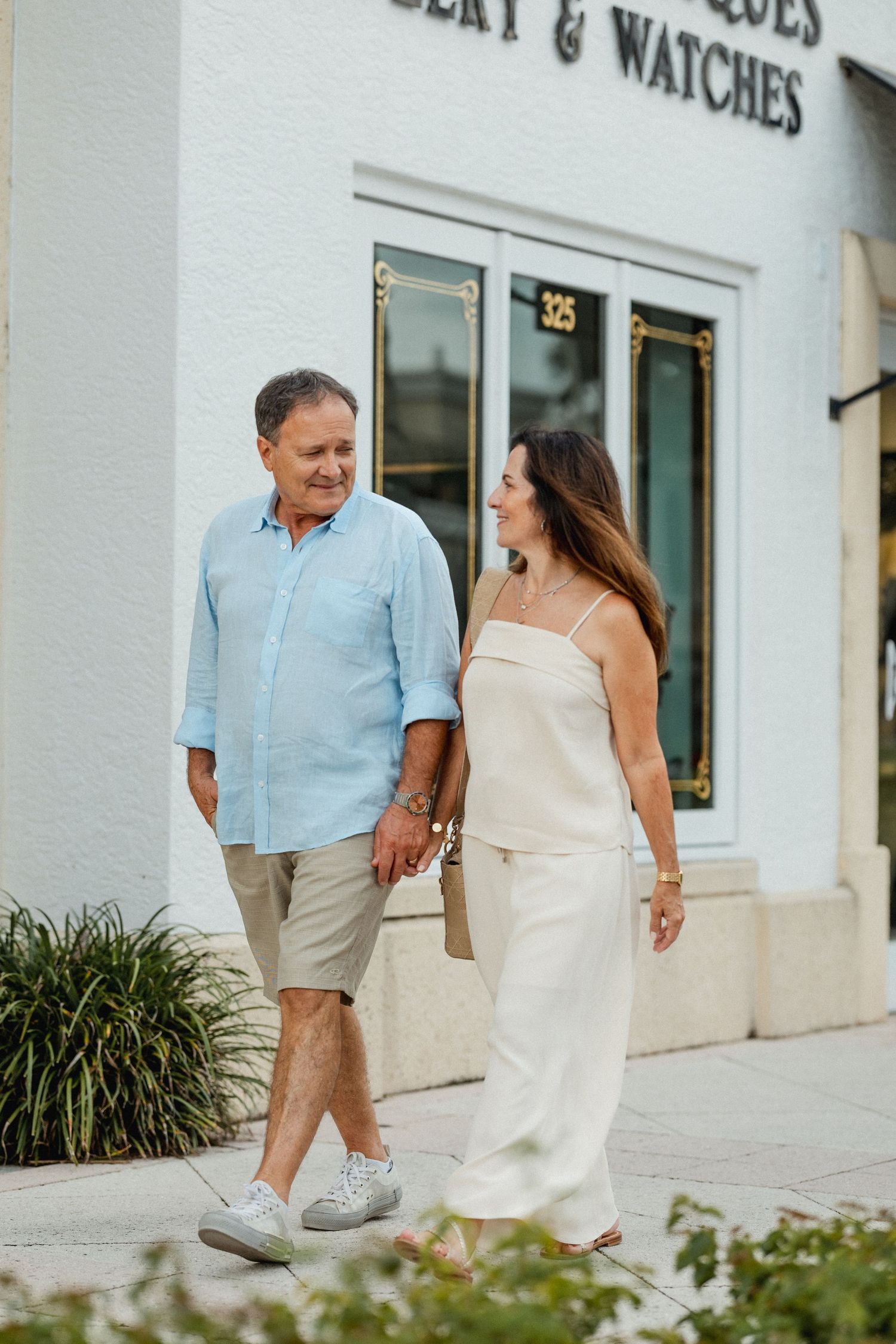 Couple walking hand in hand past a jewelry store in casual summer attire.