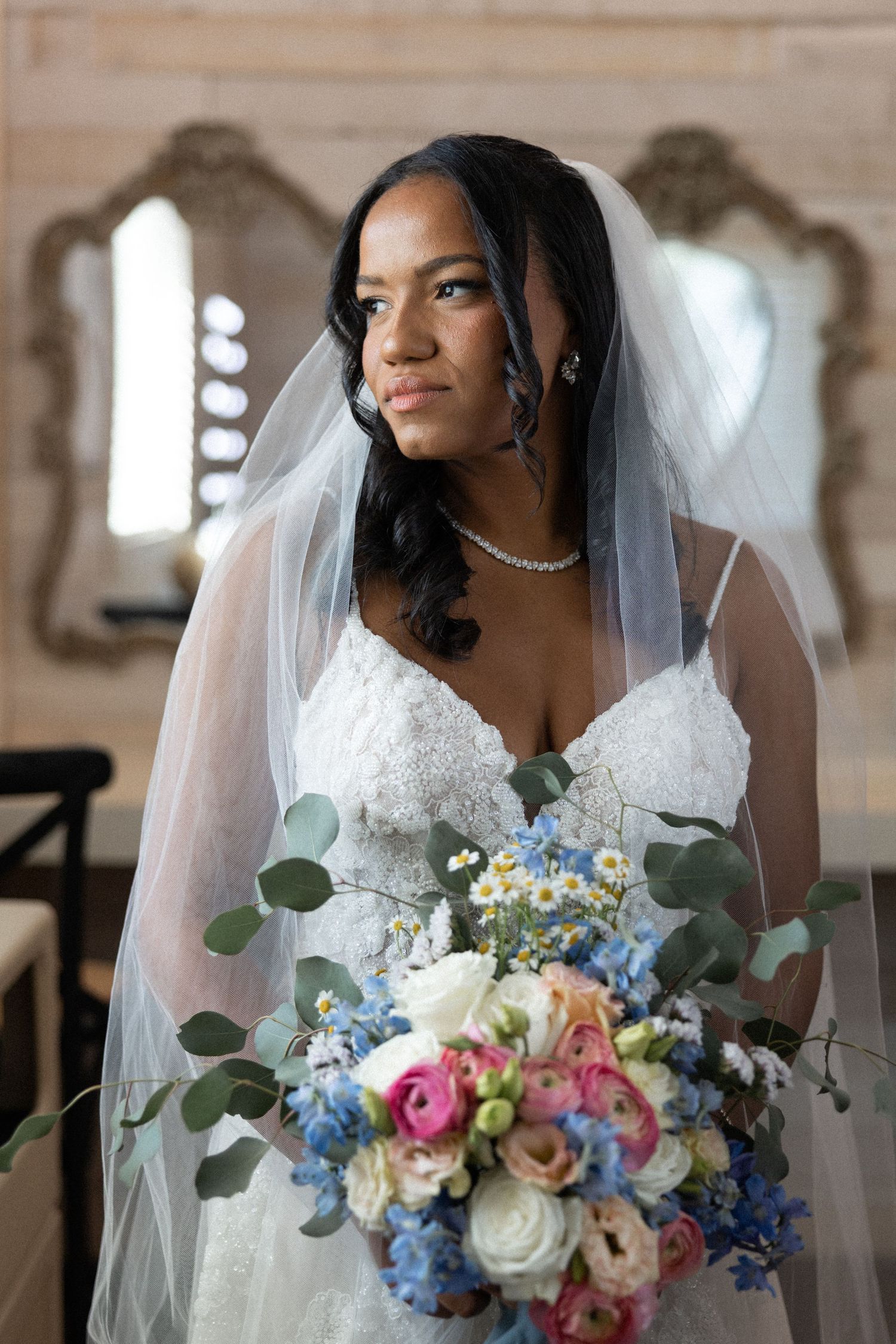 A bride in a white lace gown holds a vibrant bouquet of blue, pink and white flowers with eucalyptus greenery.