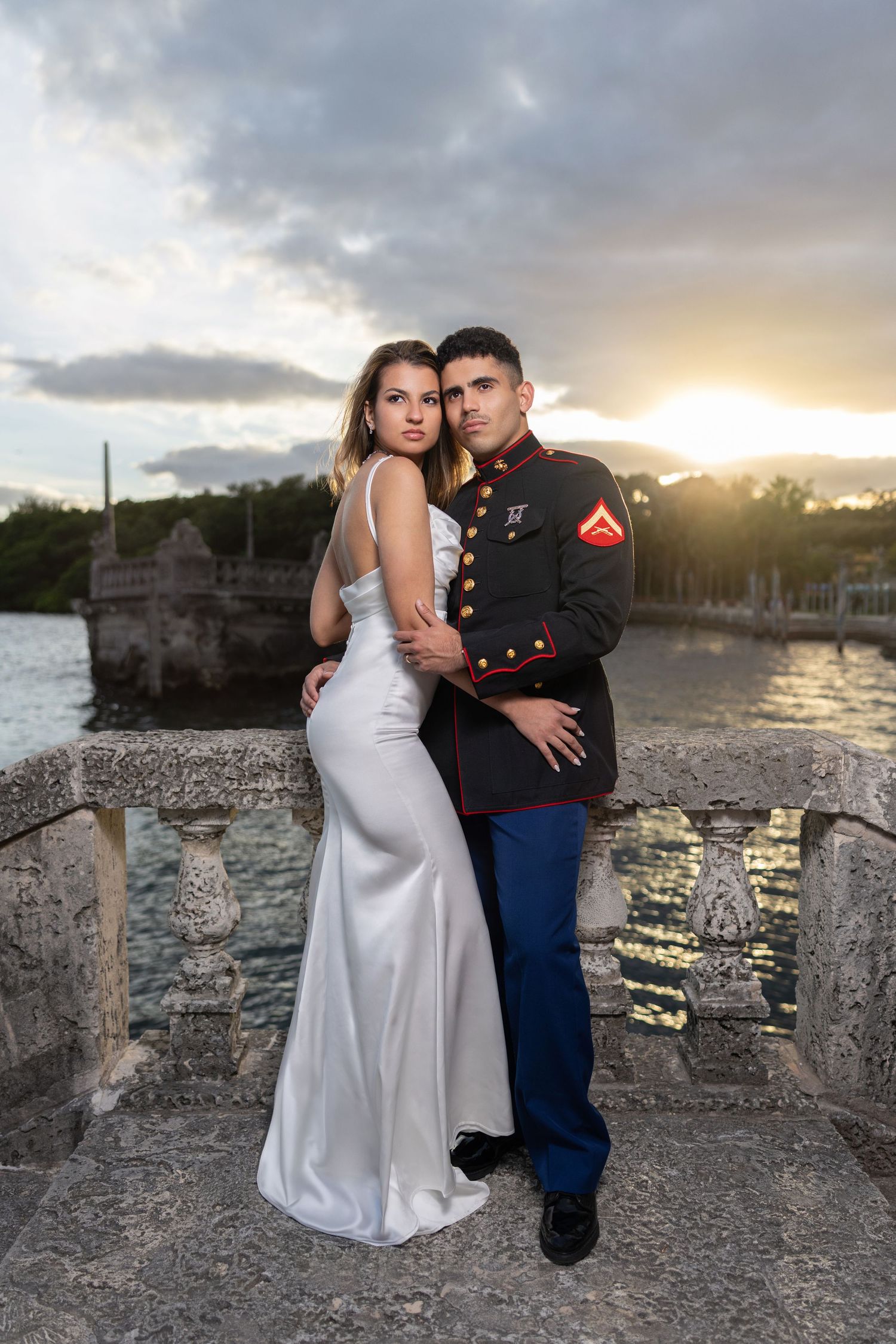 A couple in wedding attire poses at sunset on a stone wall by the water with dramatic clouds in the background.