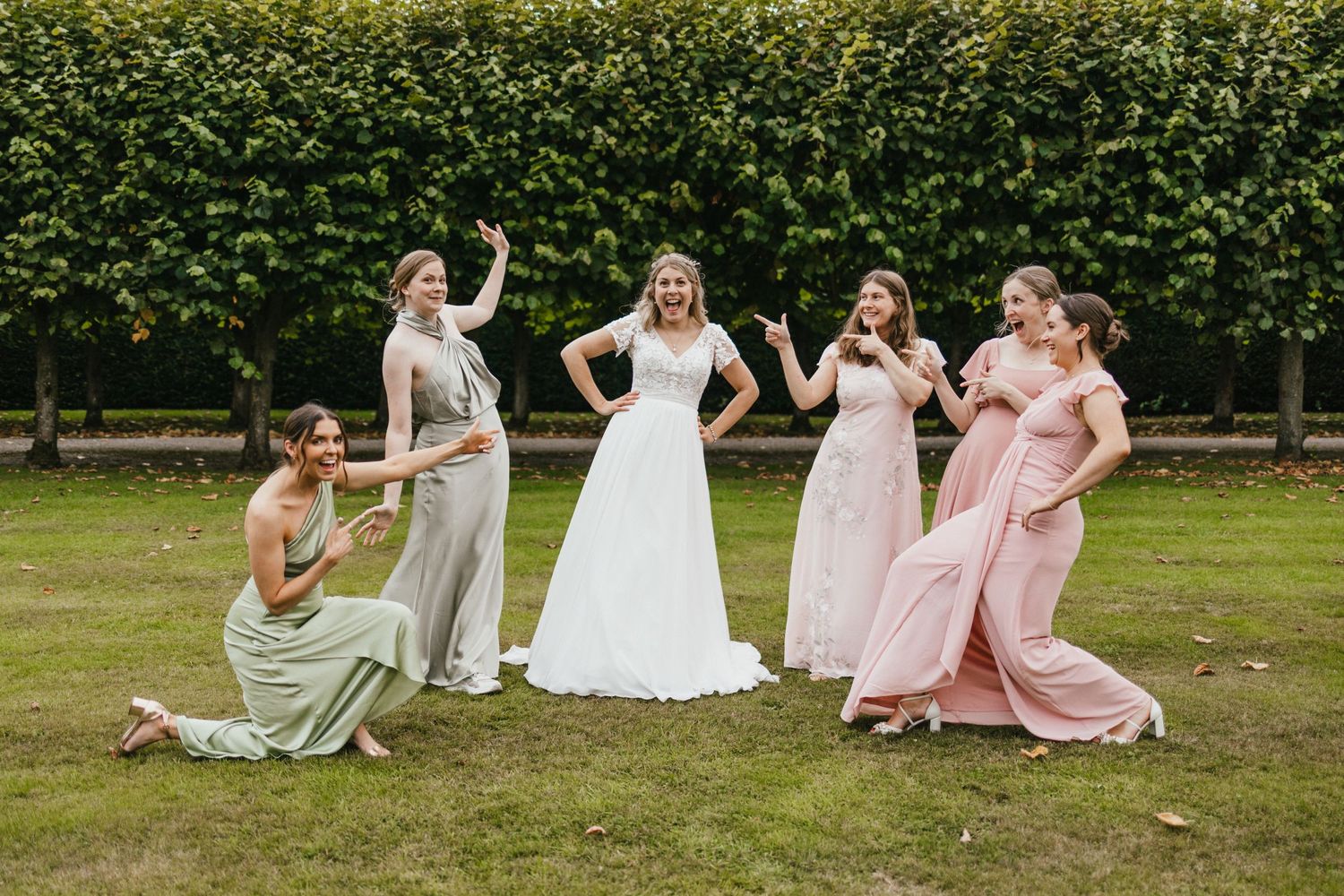 Bridesmaids in pastel dresses strike playful poses on a lawn with hedge backdrop.