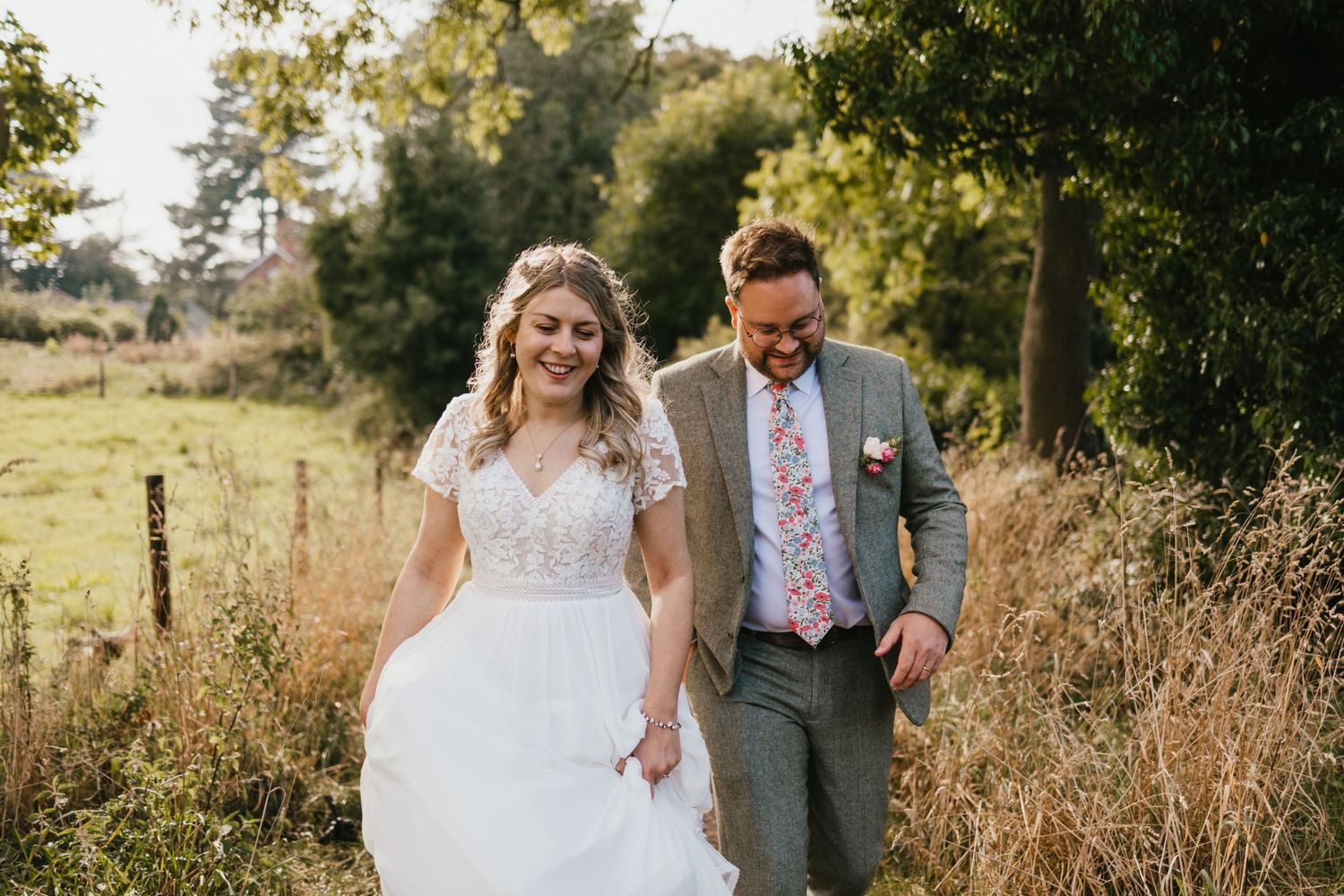 A joyful wedding couple walking together through a sunlit field with natural foliage in the background.