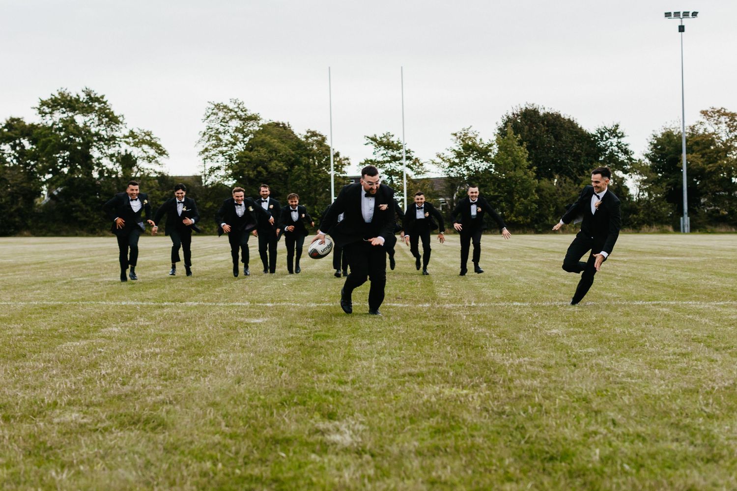 Groomsmen in black suits running in synchronized formation on sports field.