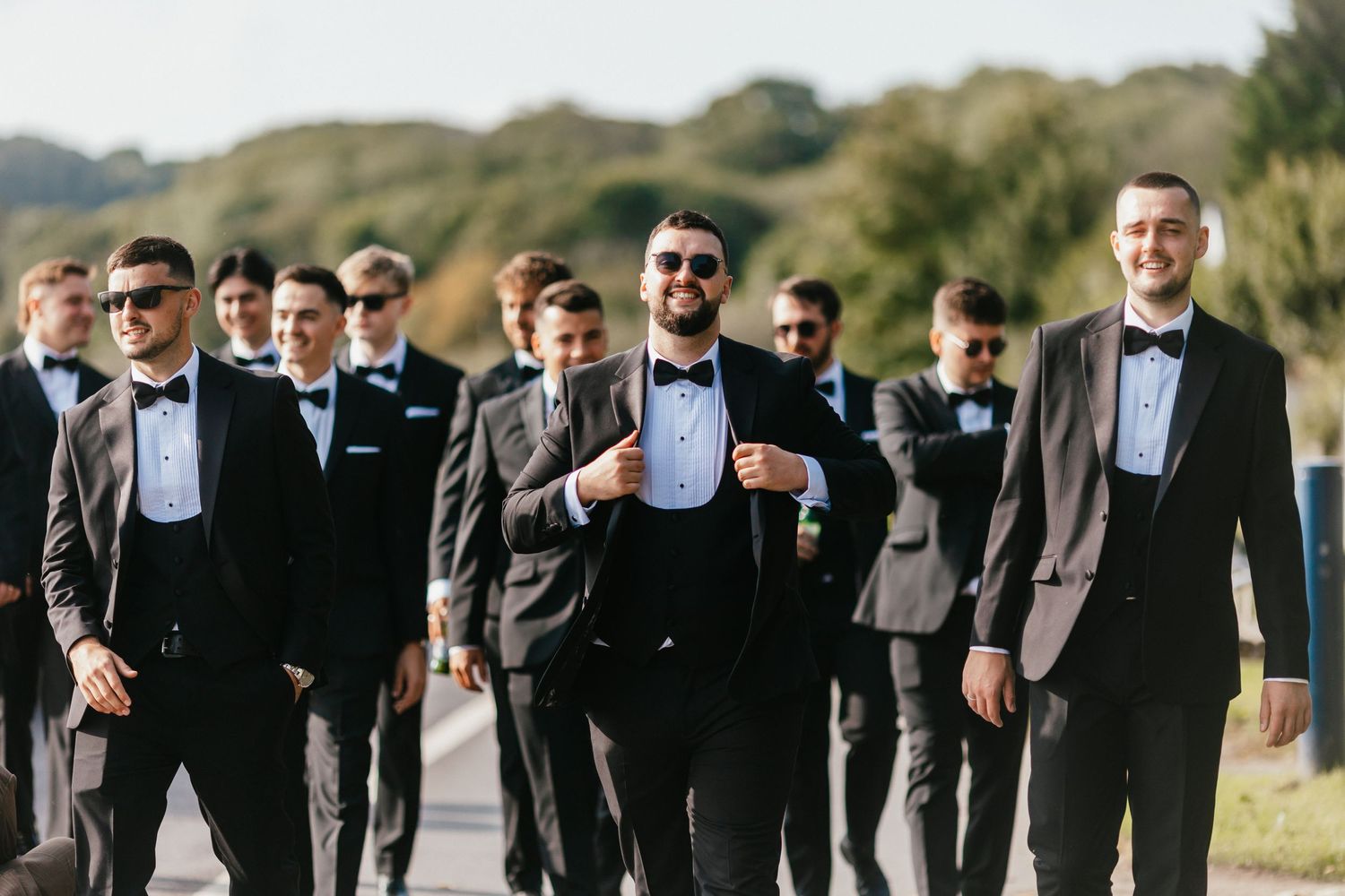 Groomsmen in black tuxedos and bow ties walking together outdoors.