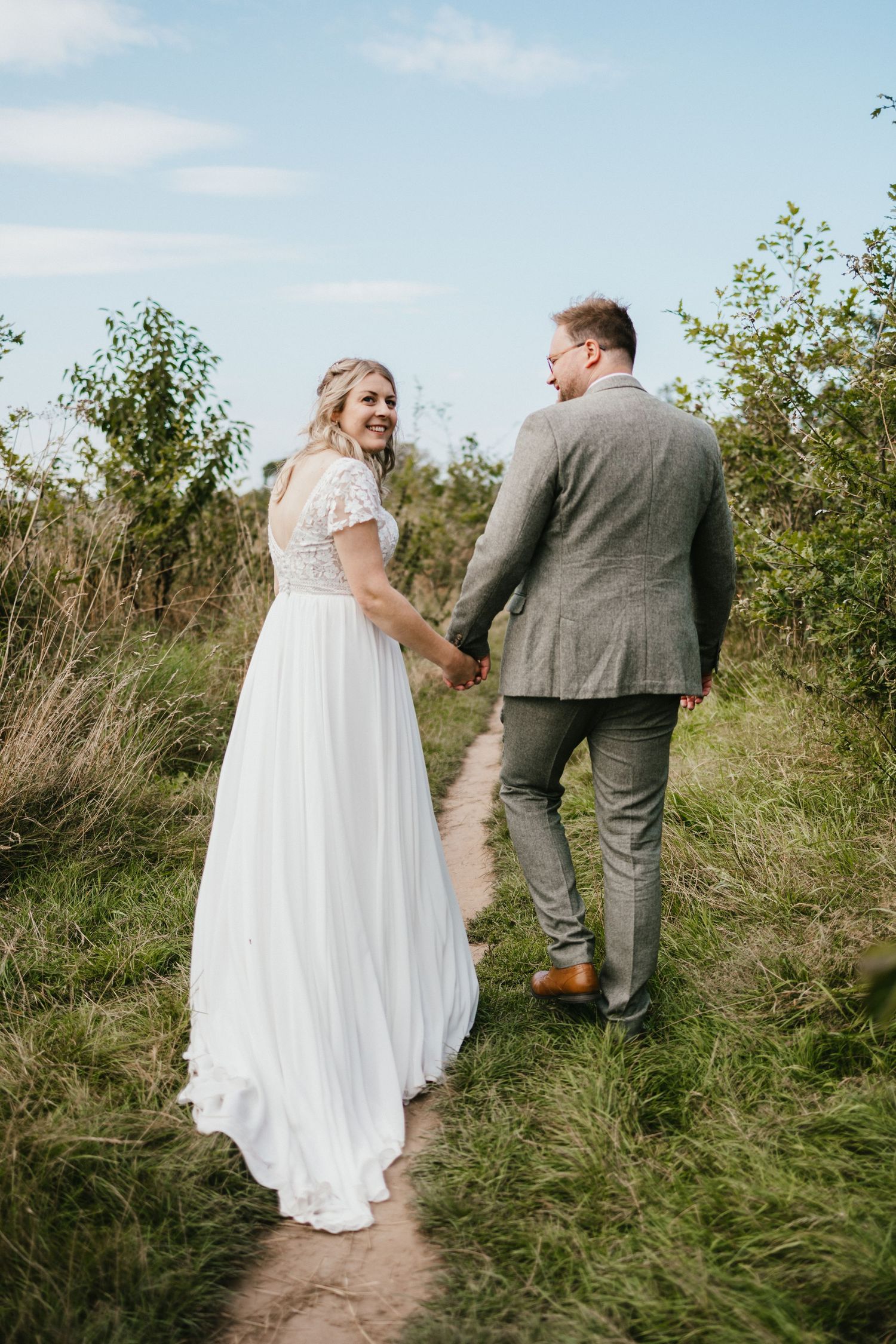 A wedding couple walking hand in hand along a nature path with flowing white dress and grey suit.