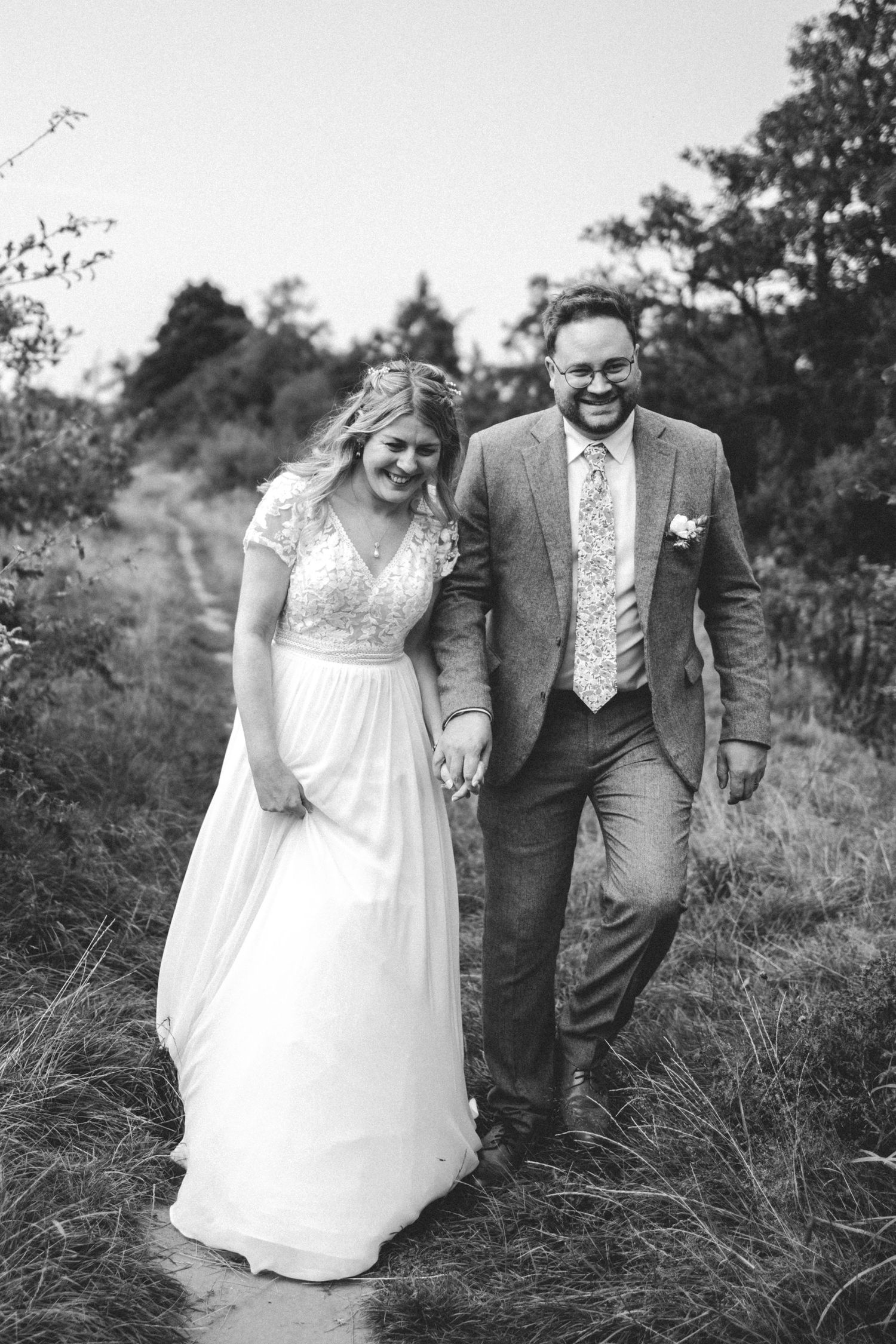 A black and white photo of newlyweds walking hand in hand down a rustic path.