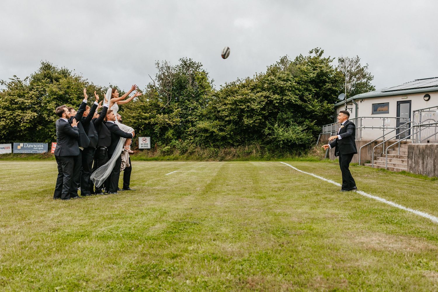 Wedding party having fun tossing rugby ball on green lawn.