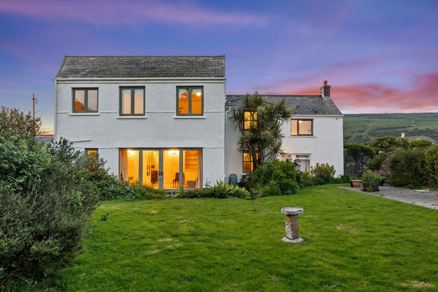 Modern house exterior with warm window lighting and green lawn at purple sunset.