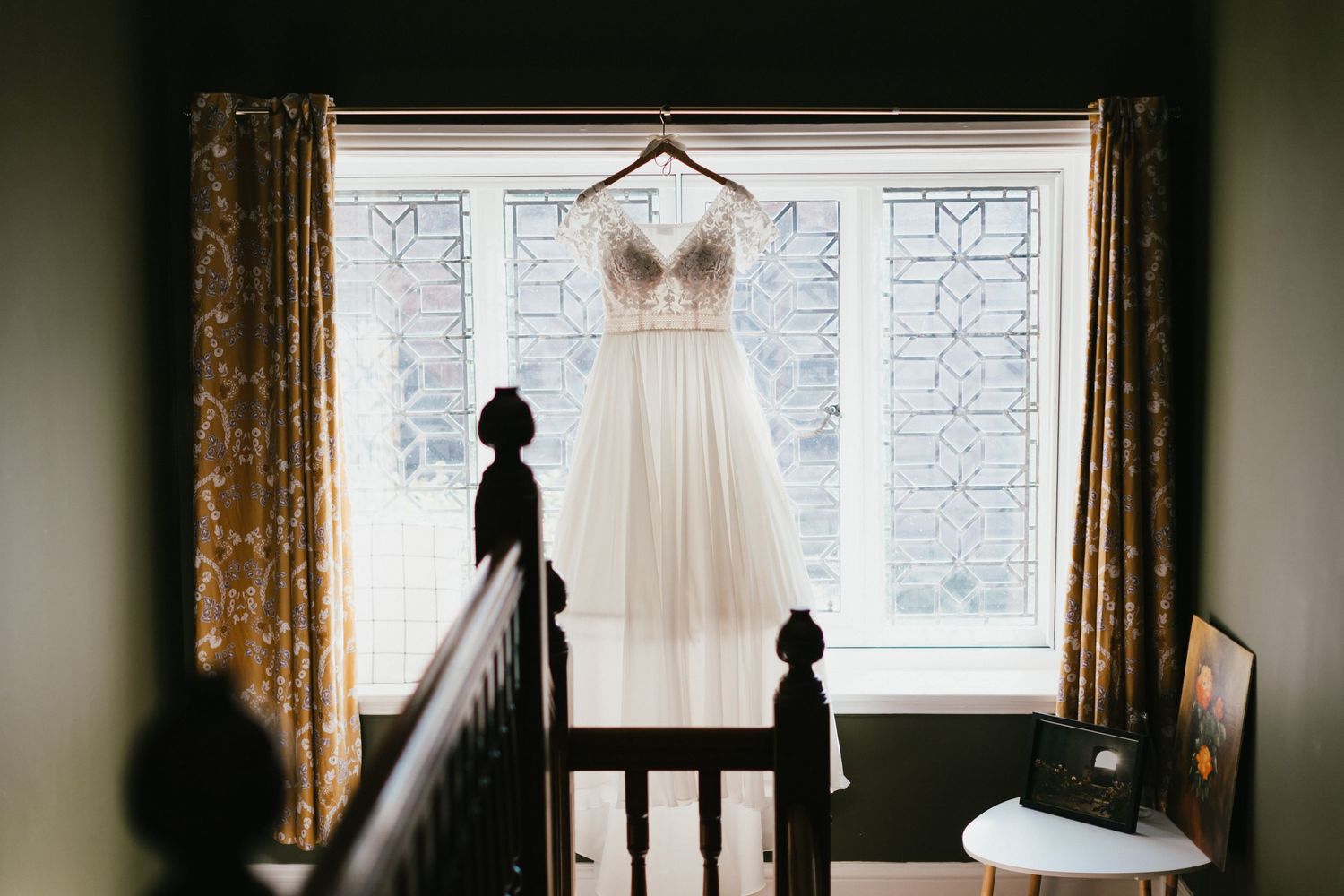 A wedding dress silhouetted in window light with ornate curtains and stair railing shadows.