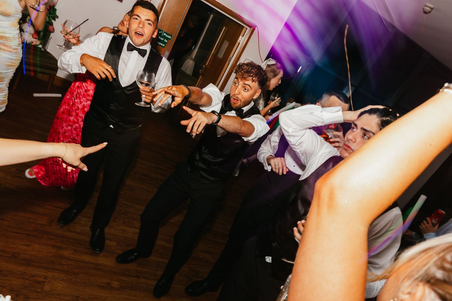 Reception guests dancing with glow sticks on a dark wooden dance floor.