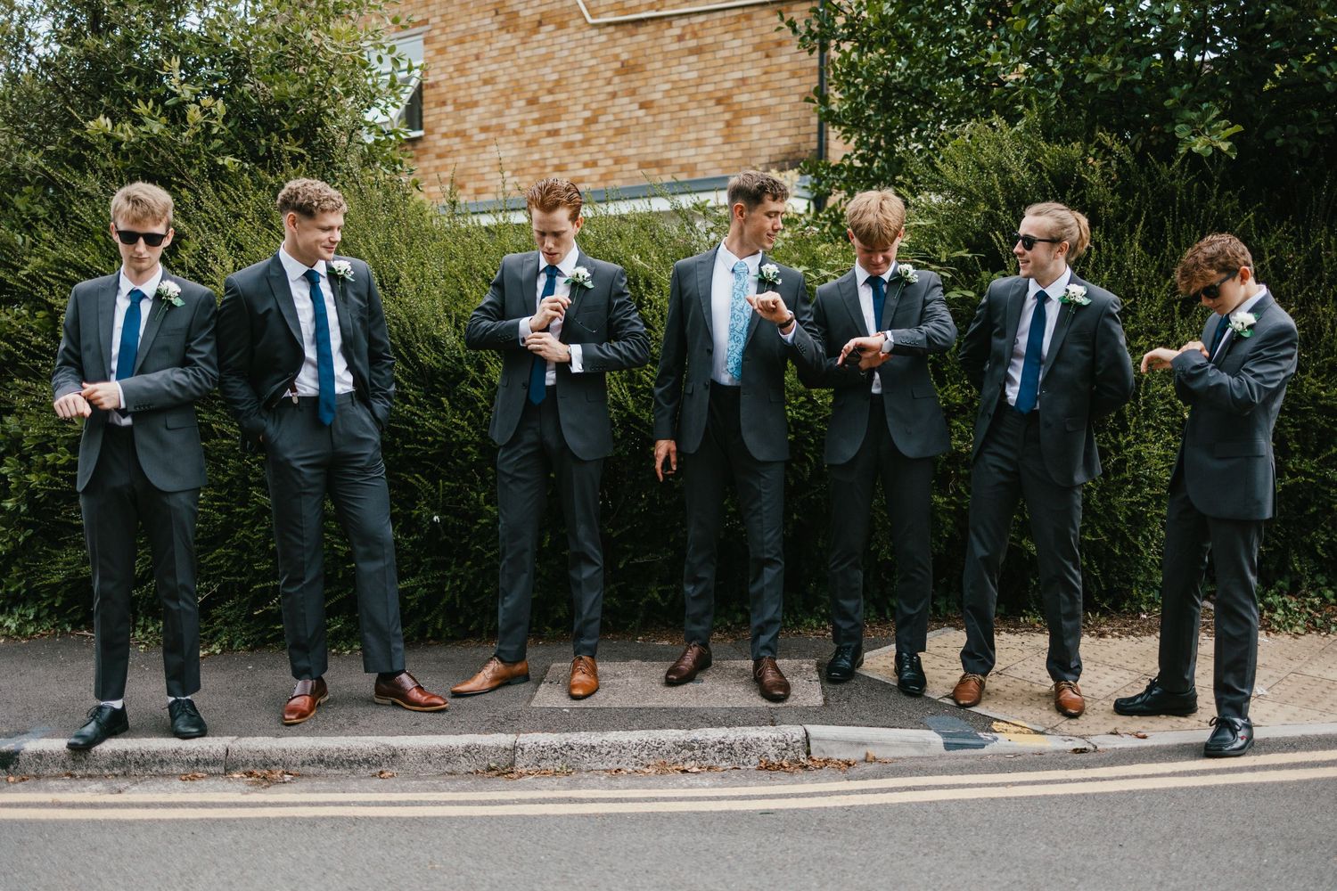 Groomsmen in dark suits checking their phones while standing in a line outdoors.