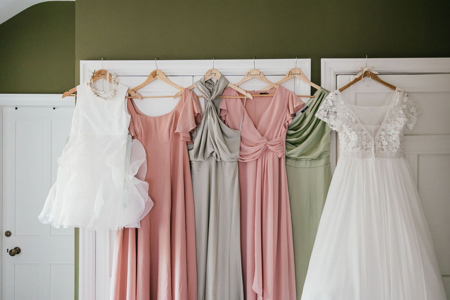 Pastel bridesmaid dresses and white wedding gowns hanging on wooden hangers against a green wall.