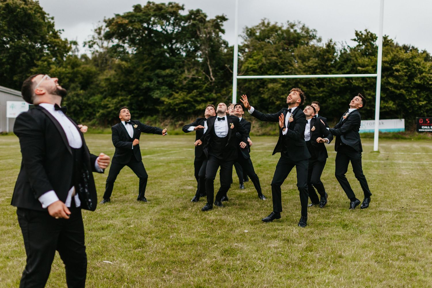 Groomsmen performing an energetic jumping celebration on a grassy field with rugby posts in the background.