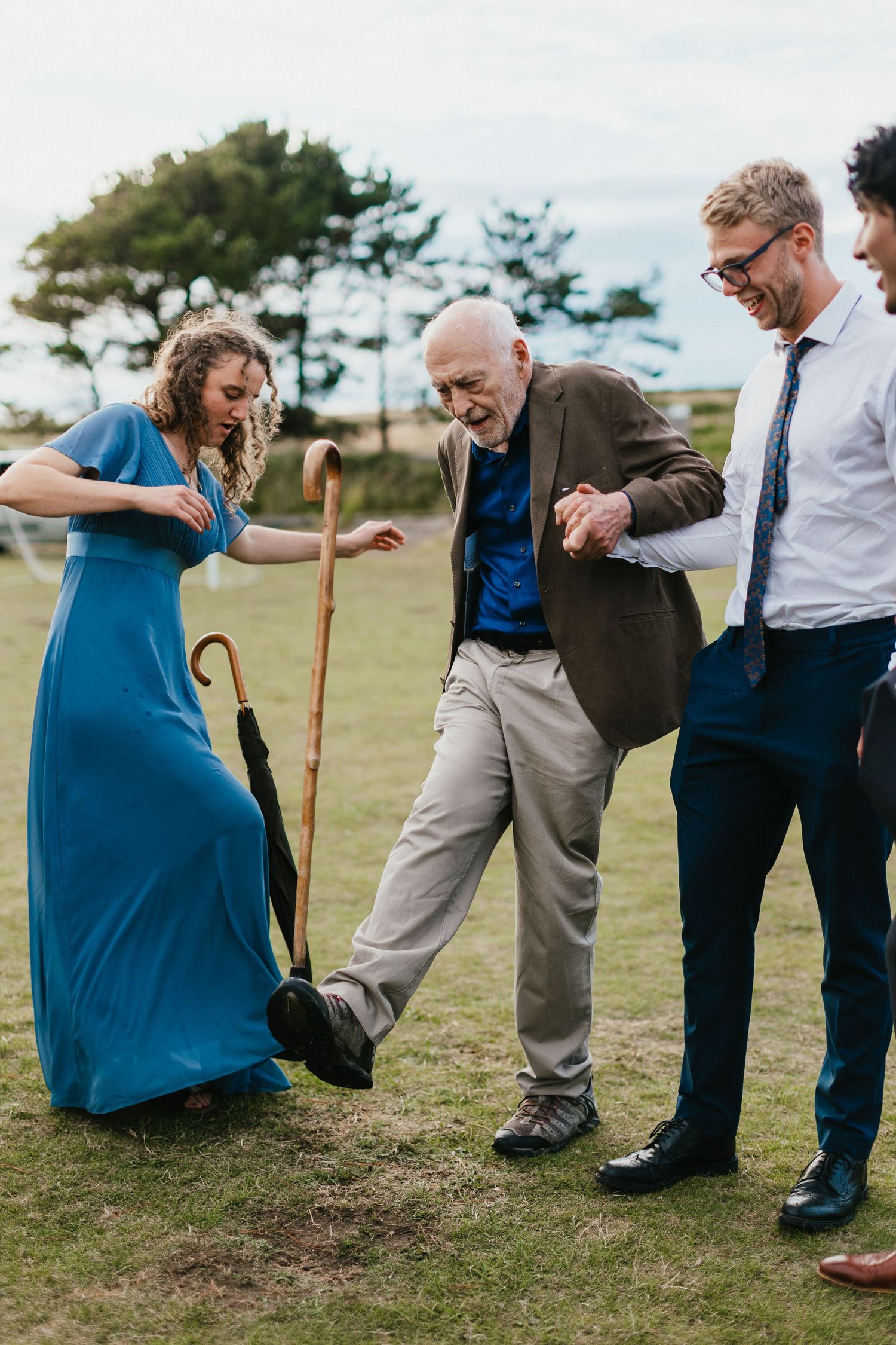 Wedding guests in formal attire dance playfully on grassy outdoor venue.