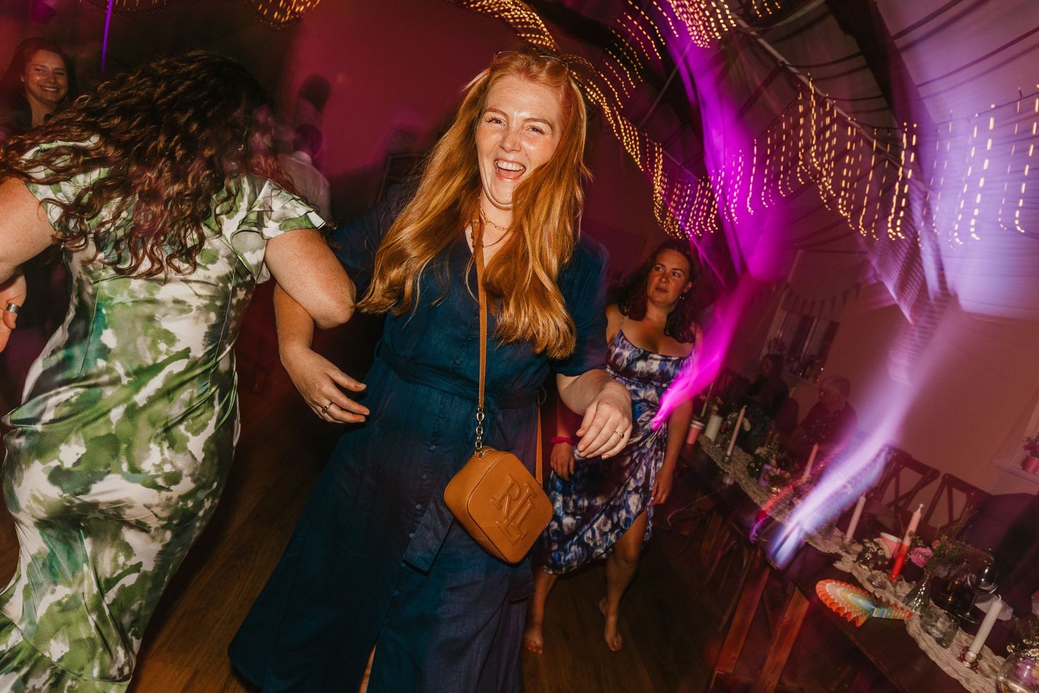 Wedding party guests dance enthusiastically under vibrant pink and purple lighting at a celebration.
