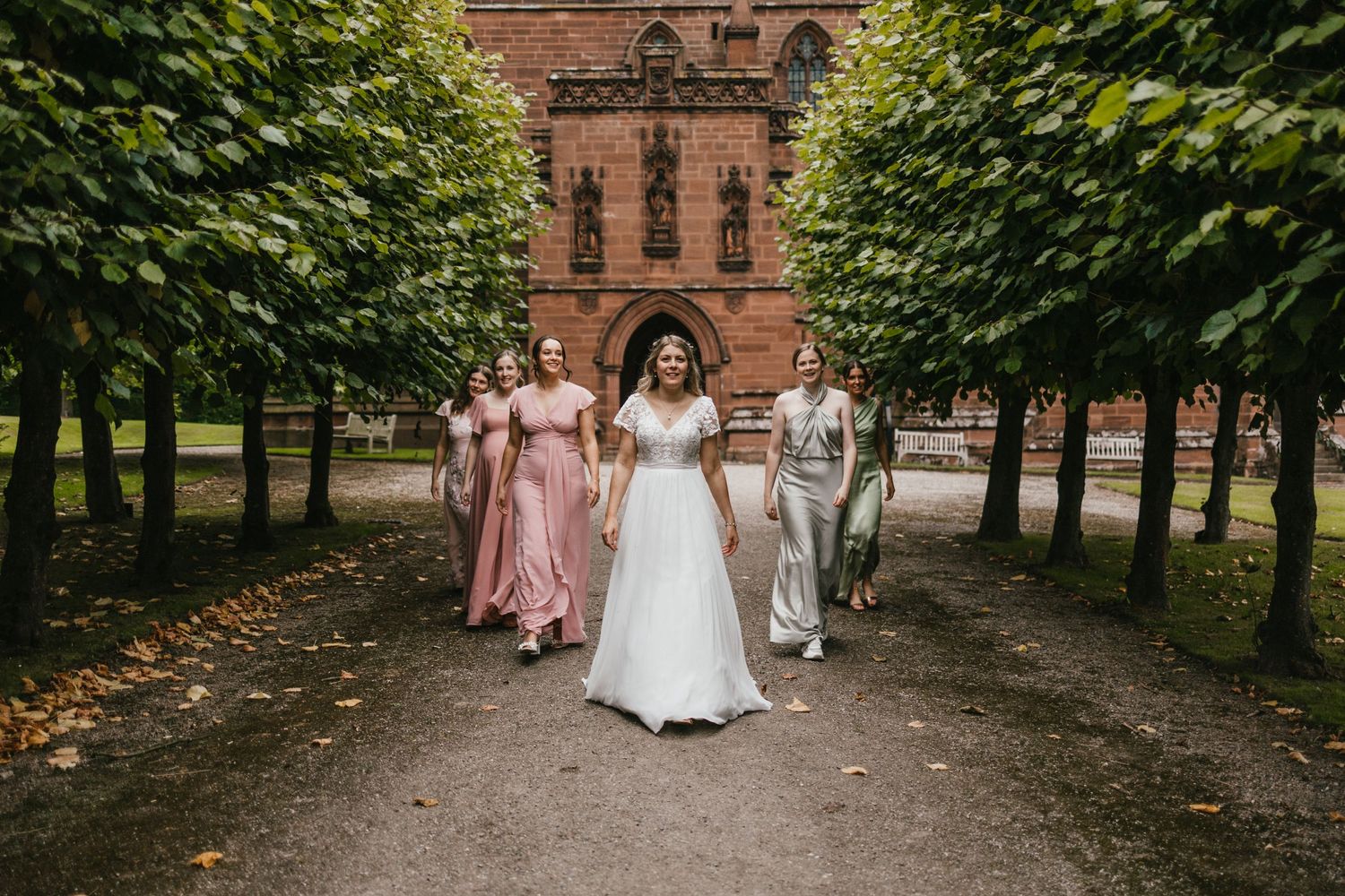 Wedding party walking down tree-lined path away from brick building.