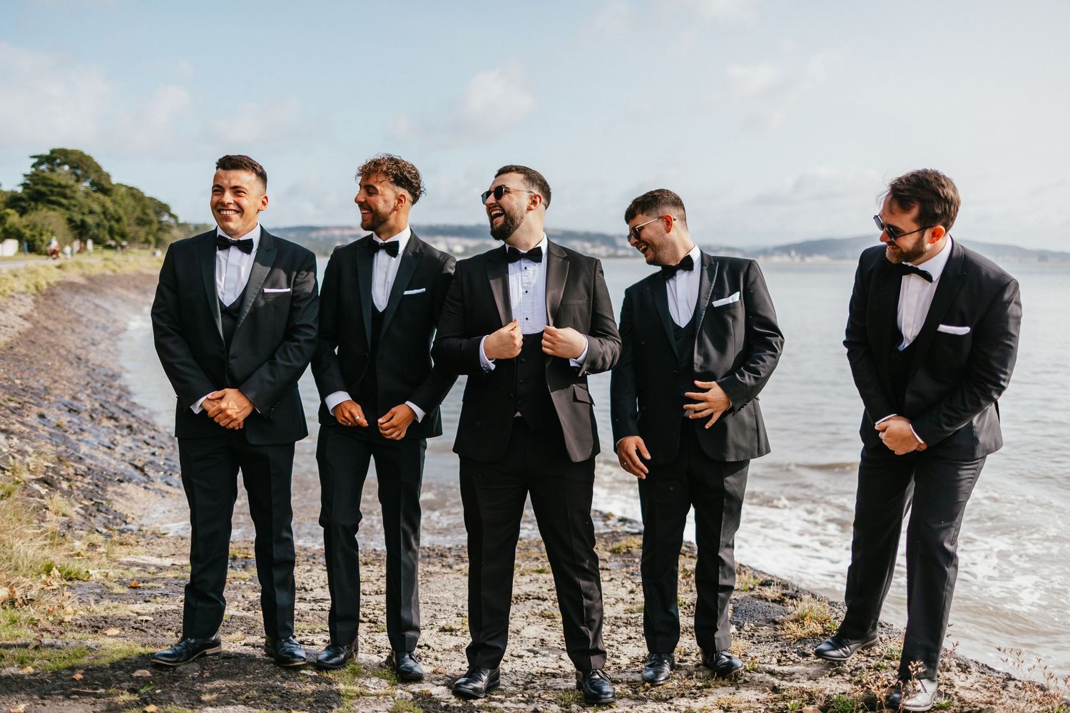 Wedding party in classic black tuxedos standing together on a beach against ocean backdrop.