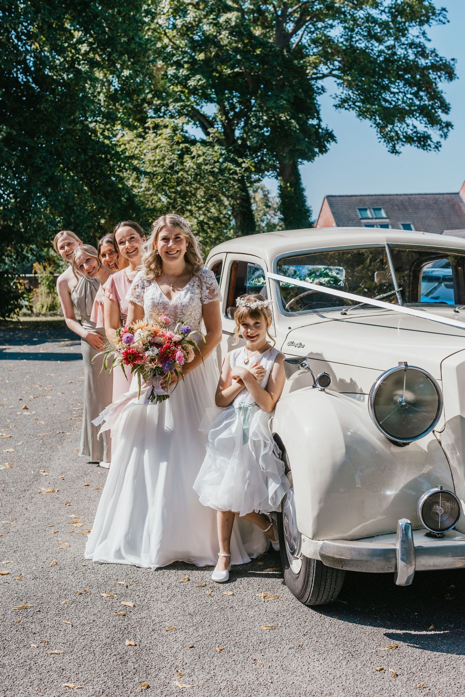 Bridal party in white dresses posing beside classic vintage car on sunny day.