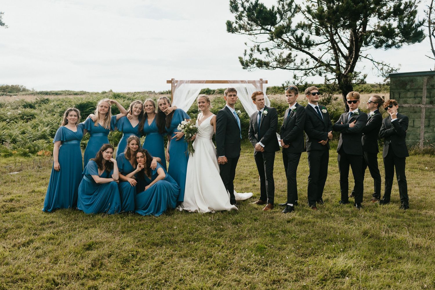 Wedding party in blue dresses and black suits posing together on a scenic hillside with white arch backdrop.