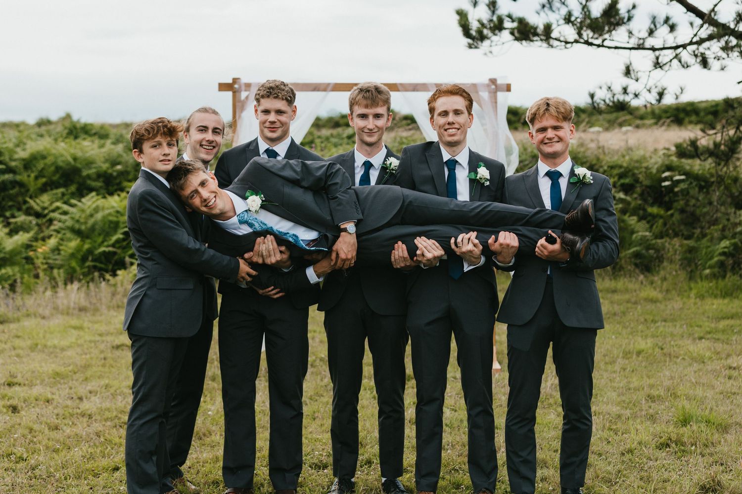 Groomsmen in dark suits playfully posing together while holding a long dark object outdoors.