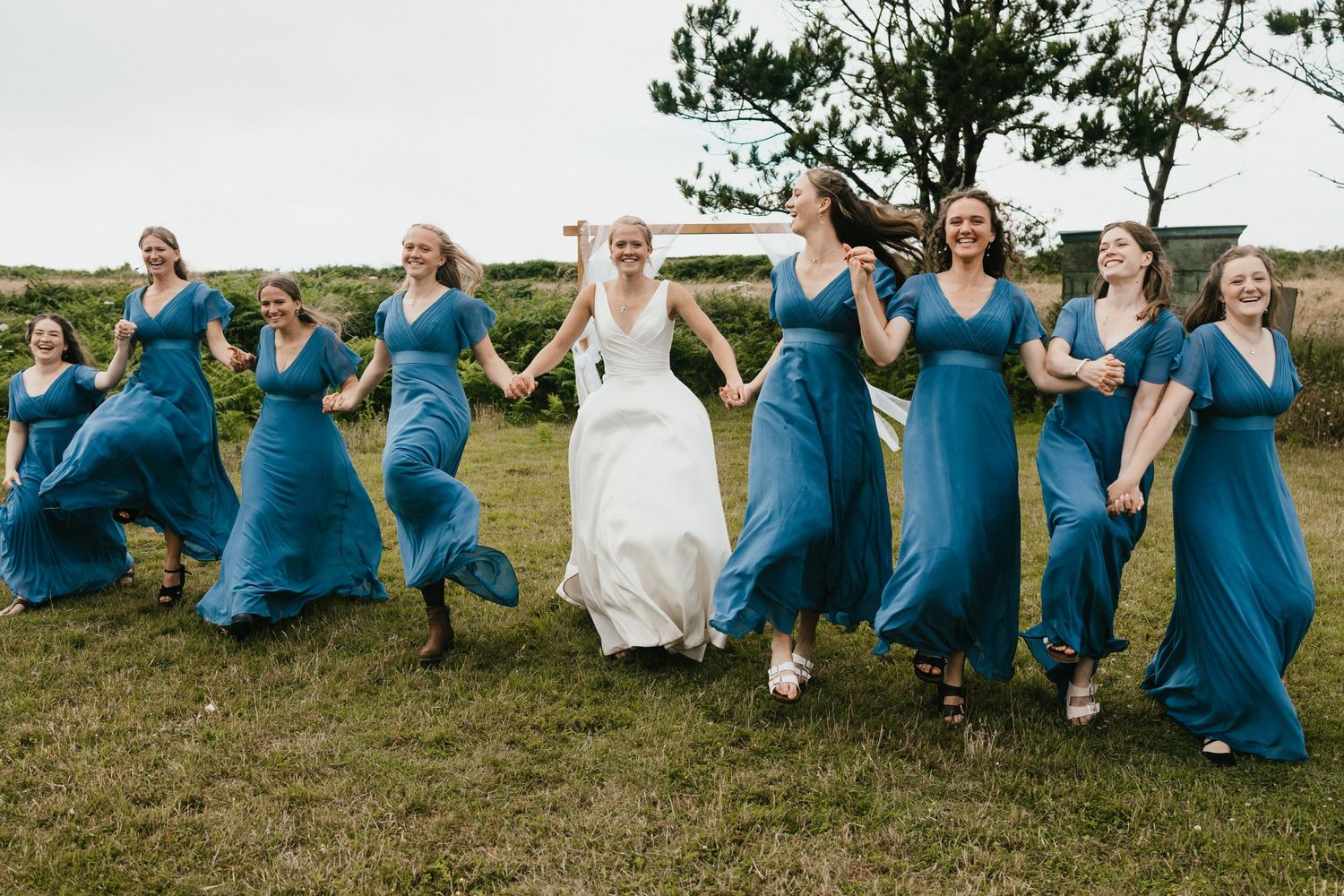 Bridesmaids in flowing blue dresses dancing with bride across grass in coordinated outdoor photo.