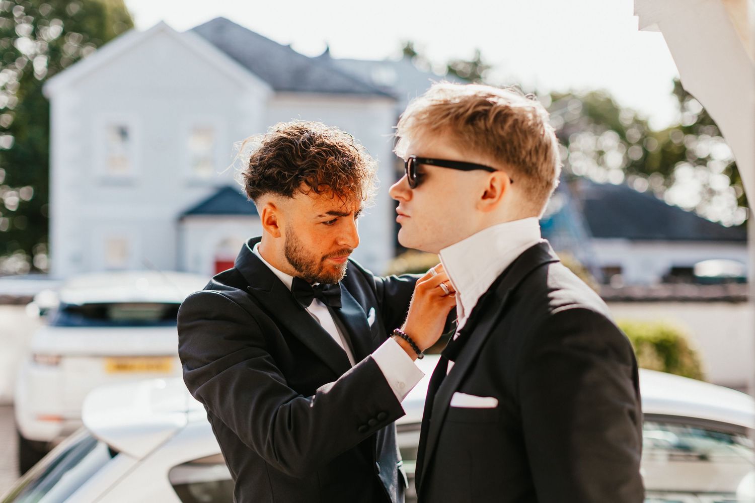 Two groomsmen in black tuxedos adjusting bow ties in bright sunlight.