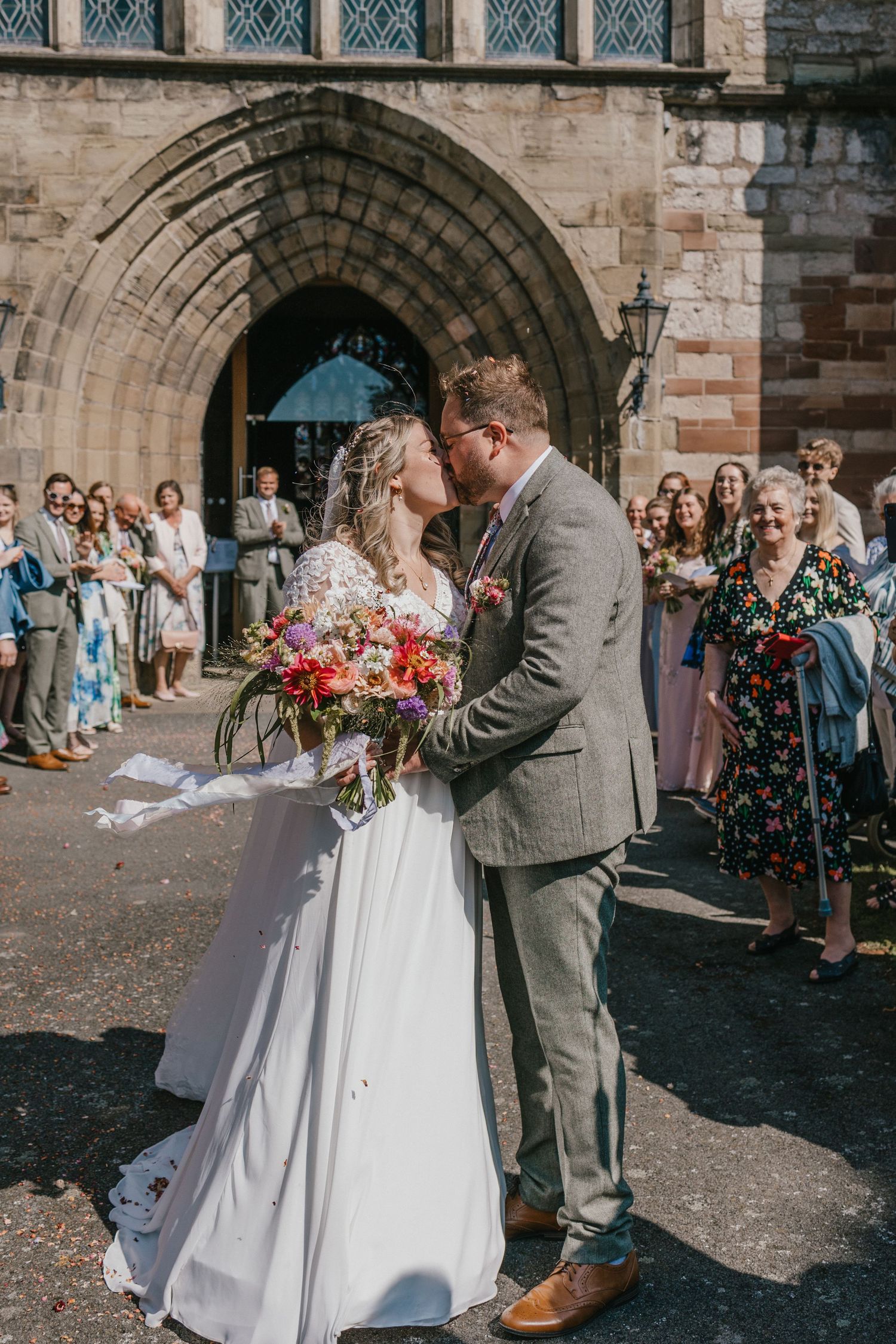Wedding ceremony outside historic stone church with guests watching the newlyweds share their first kiss.