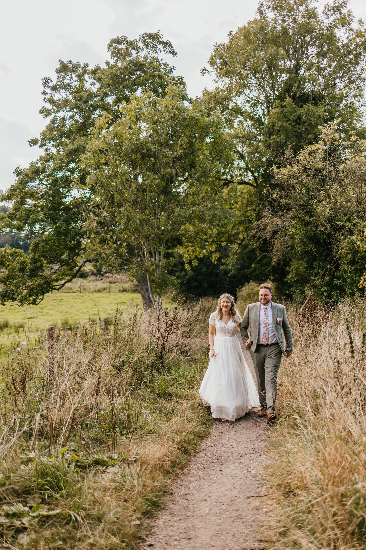 Newlyweds walking hand in hand along a nature trail with tall trees in the background.