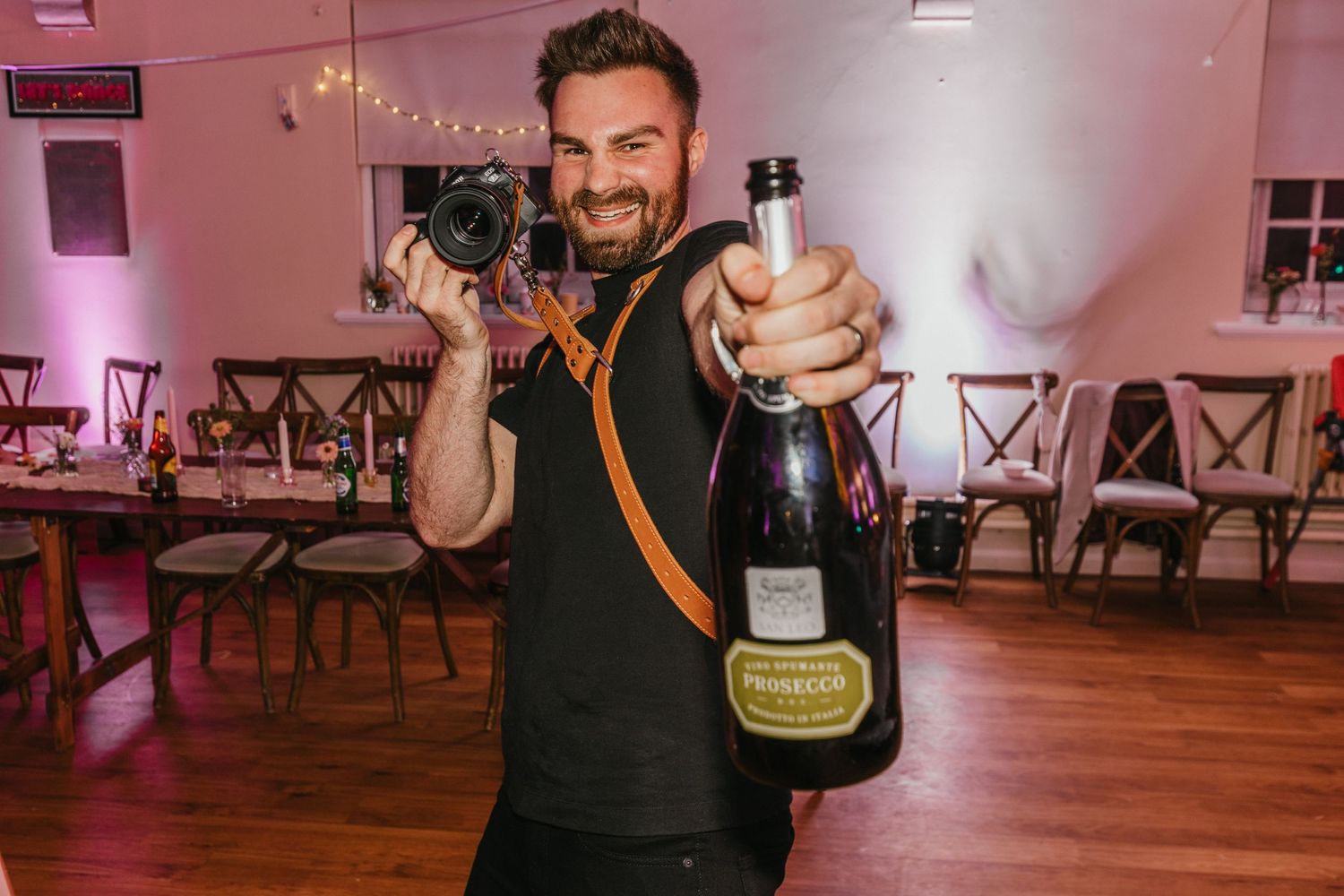 Person in black shirt holding champagne bottles in a pink-lit room with wooden floors.
