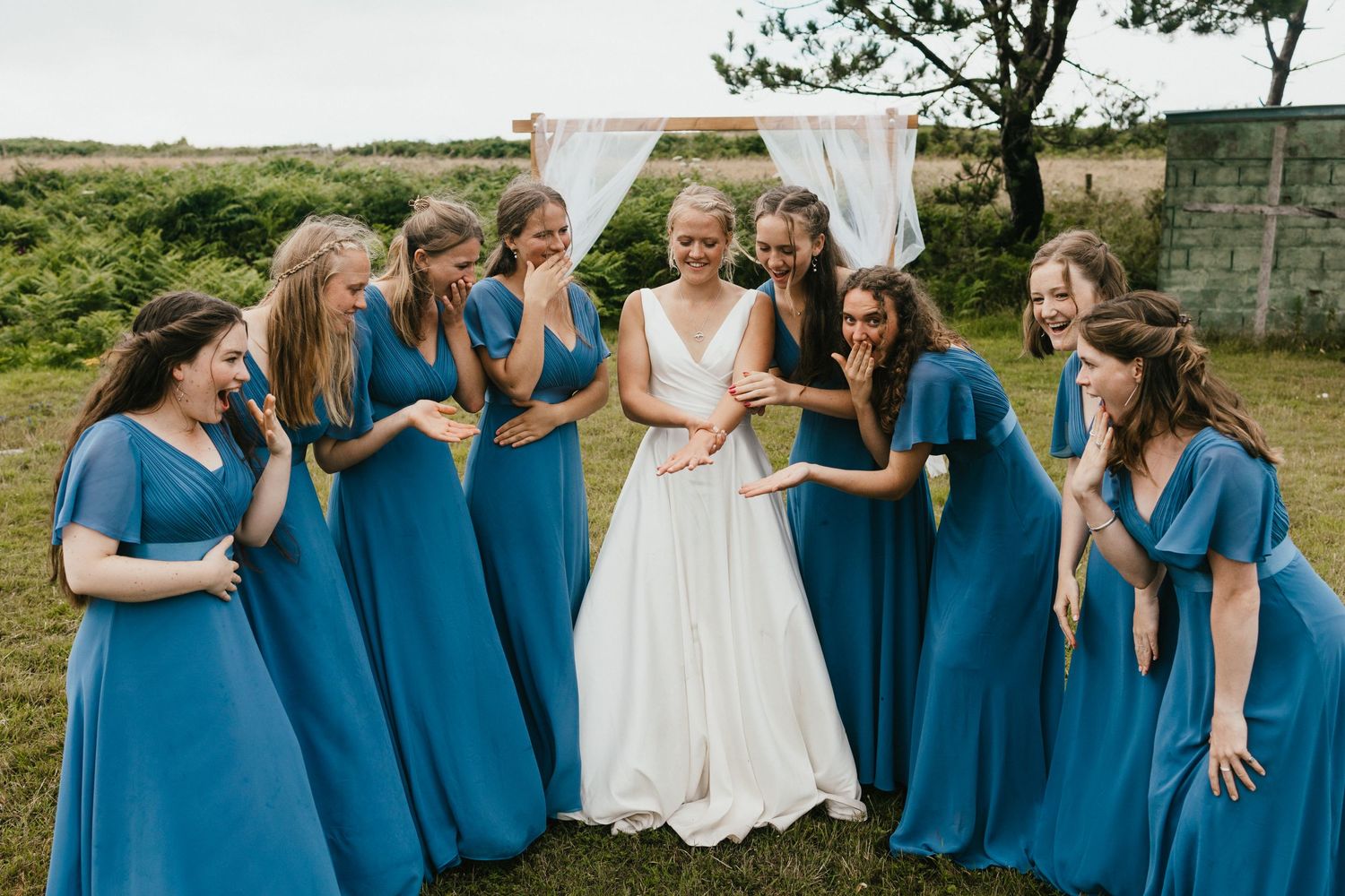 Bridesmaids in matching blue dresses gather around bride in white gown for candid outdoor photo.