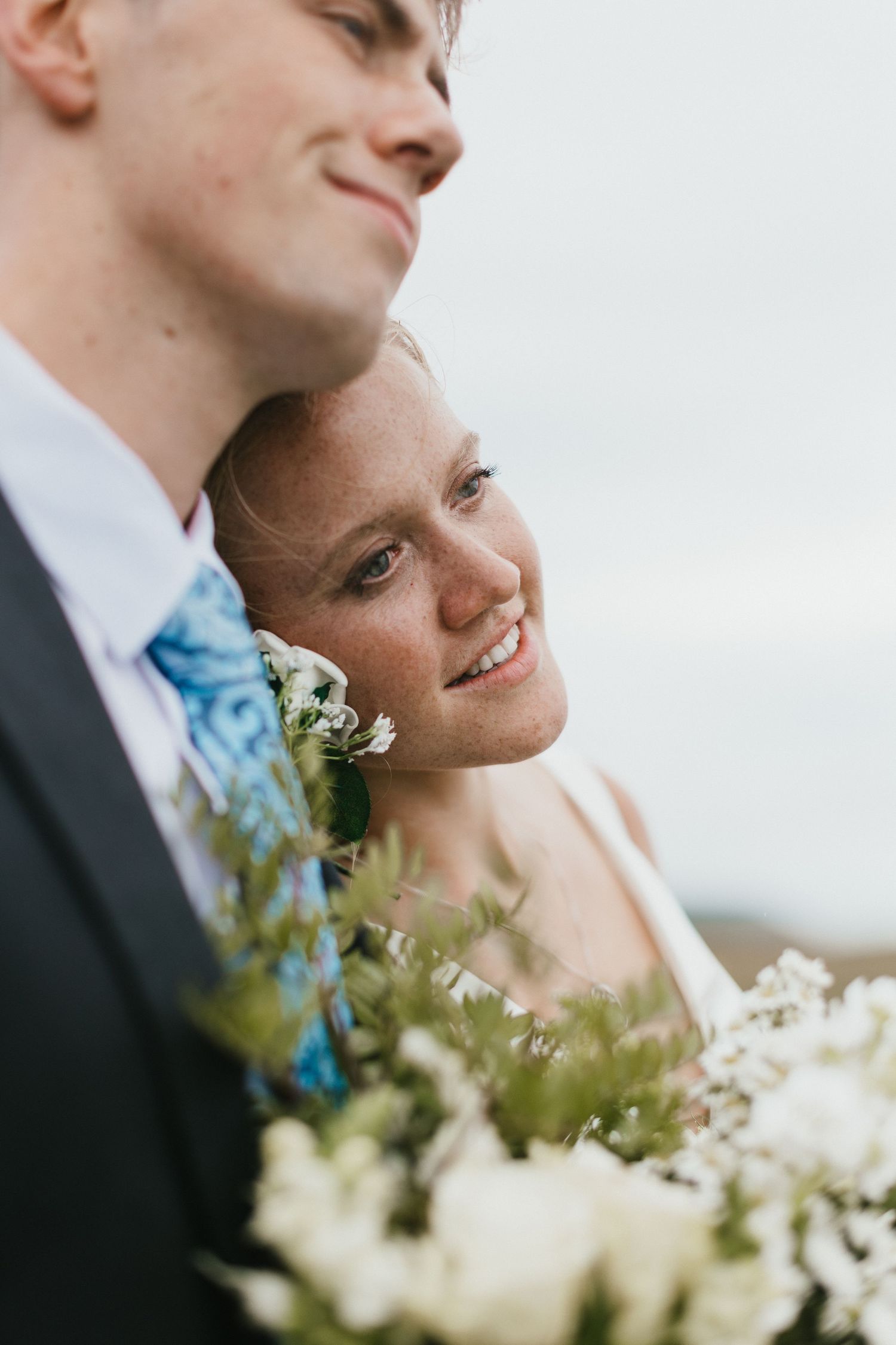 A tender wedding moment captured against an overcast sky with white flowers in the foreground.