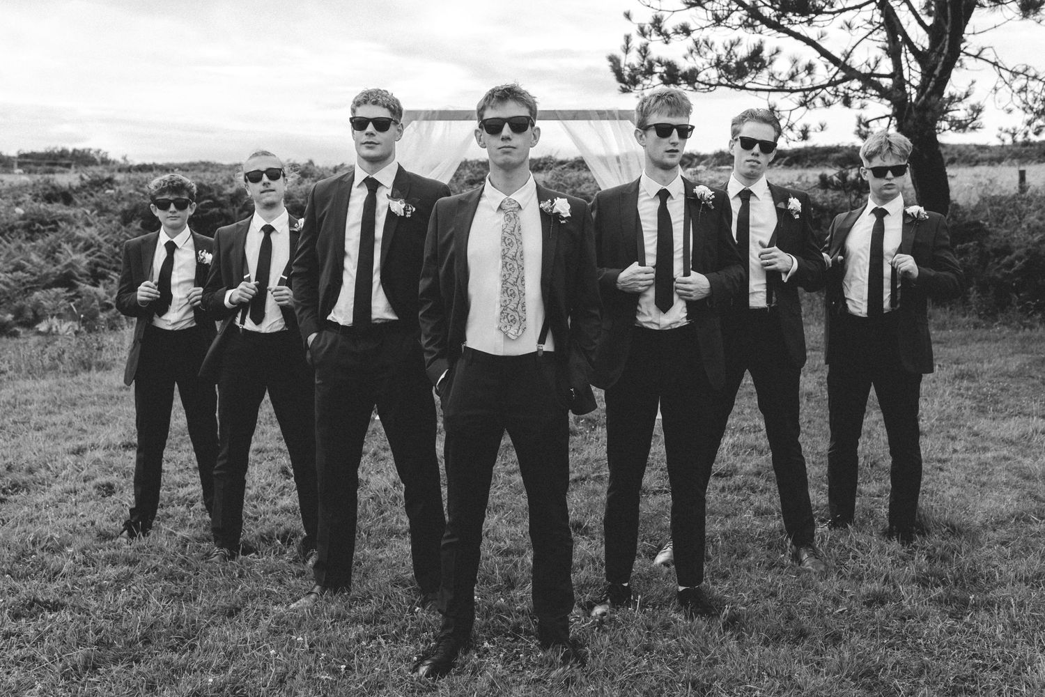Group of groomsmen in black suits and sunglasses posing dramatically in black and white photo.