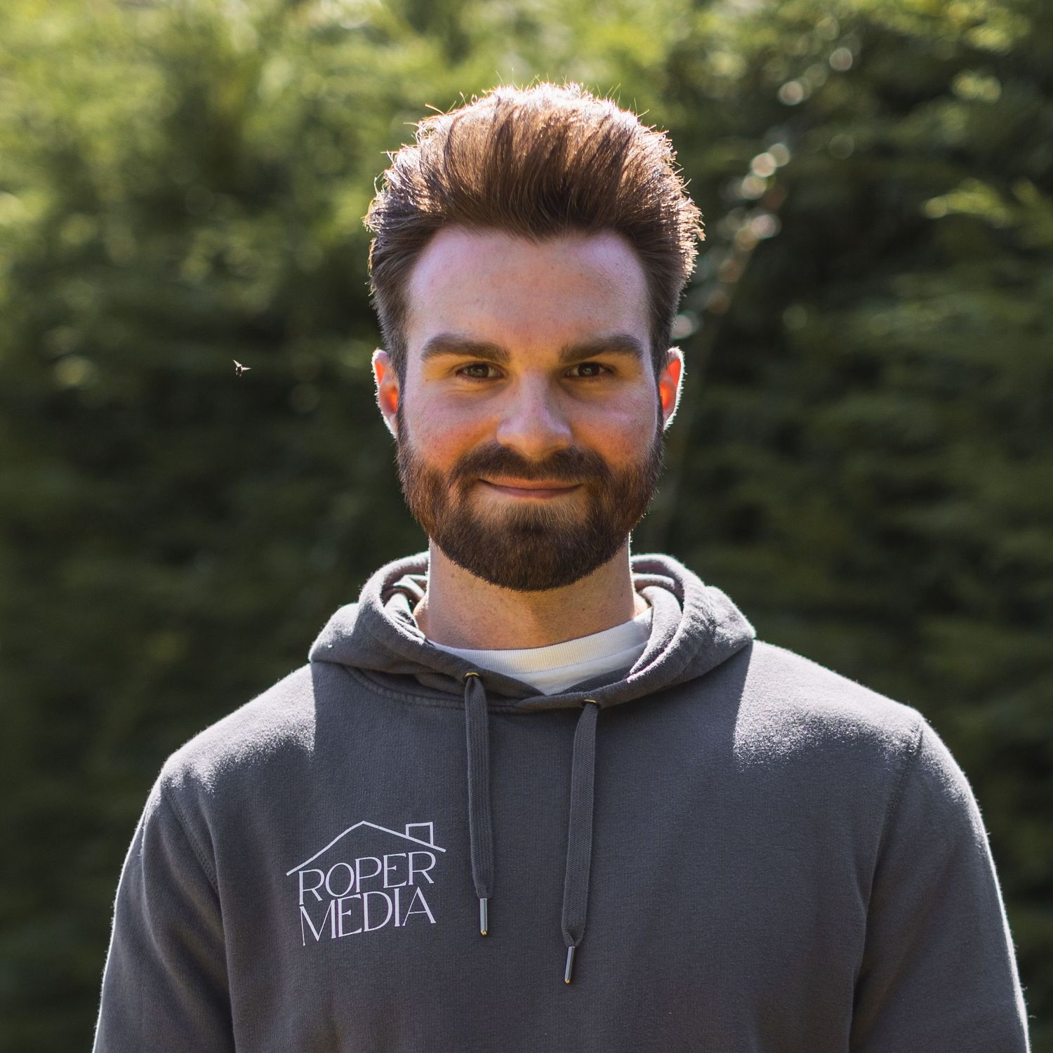 Person wearing a grey hoodie with a business logo smiling outdoors.