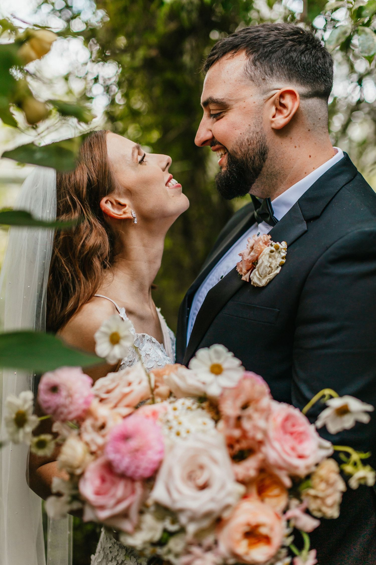 A romantic wedding portrait of a couple sharing a joyful laugh with a beautiful pink and peach floral bouquet.