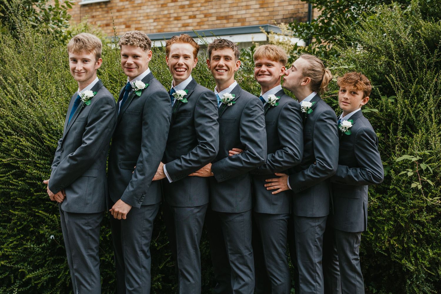 Wedding party in matching navy suits pose in a line against greenery backdrop.