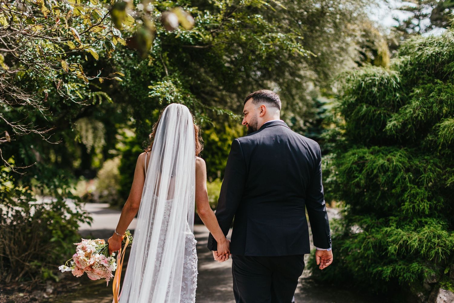 Newlyweds walking through a garden path holding pink floral bouquets.