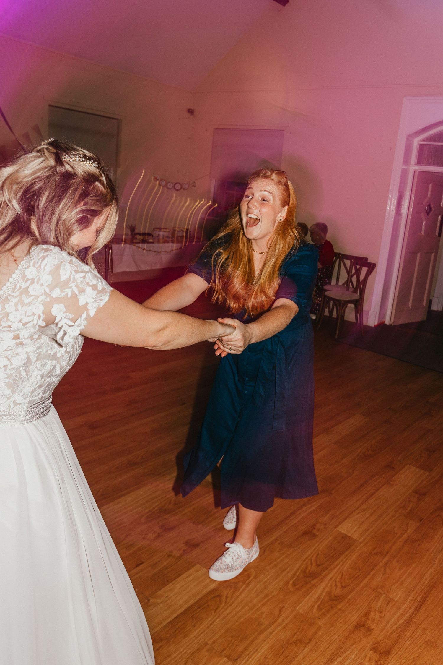 Two guests dance together on a wooden floor under pink lighting at a wedding celebration.