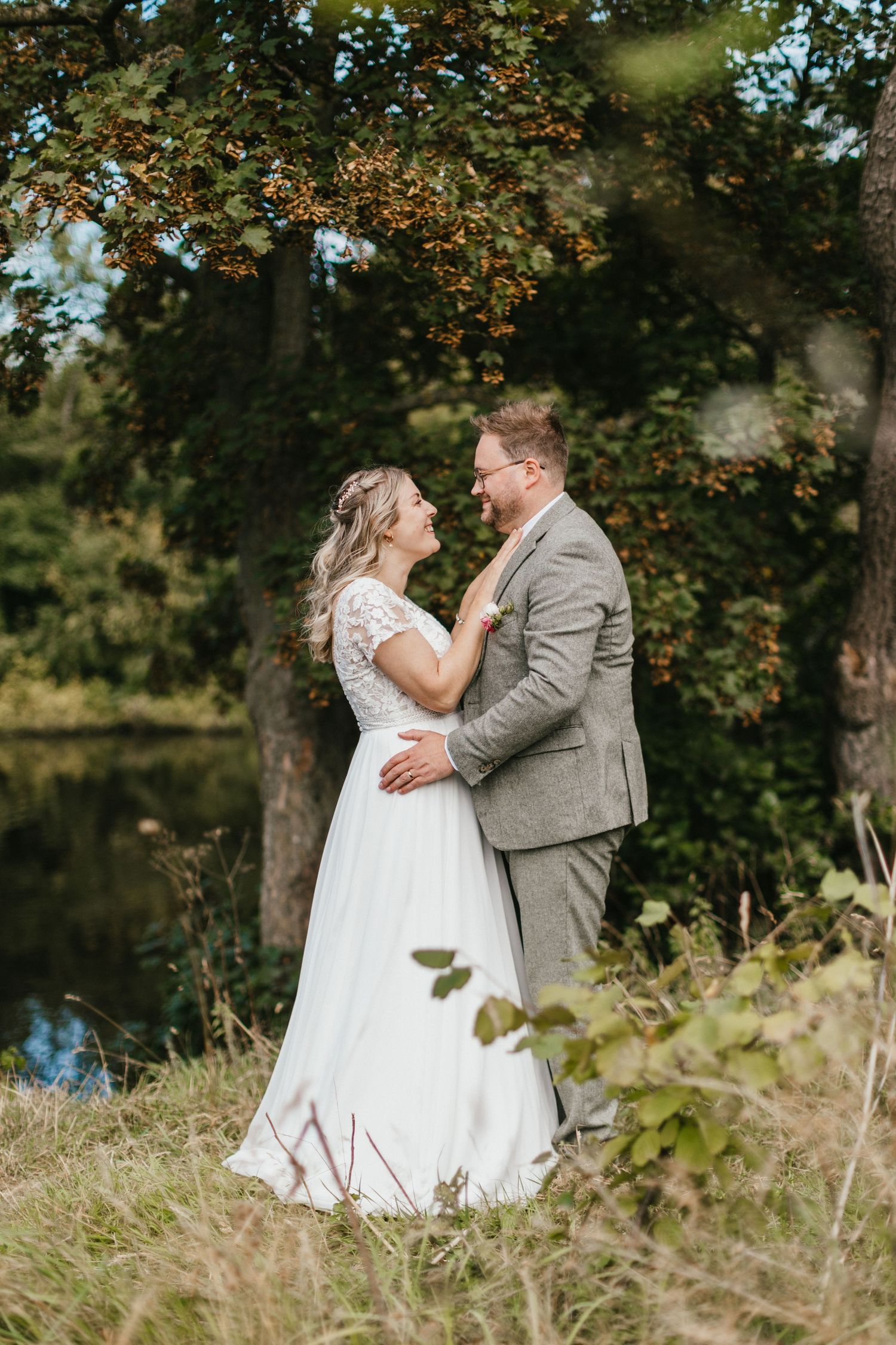 A wedding couple shares an intimate moment by a pond surrounded by autumn foliage.