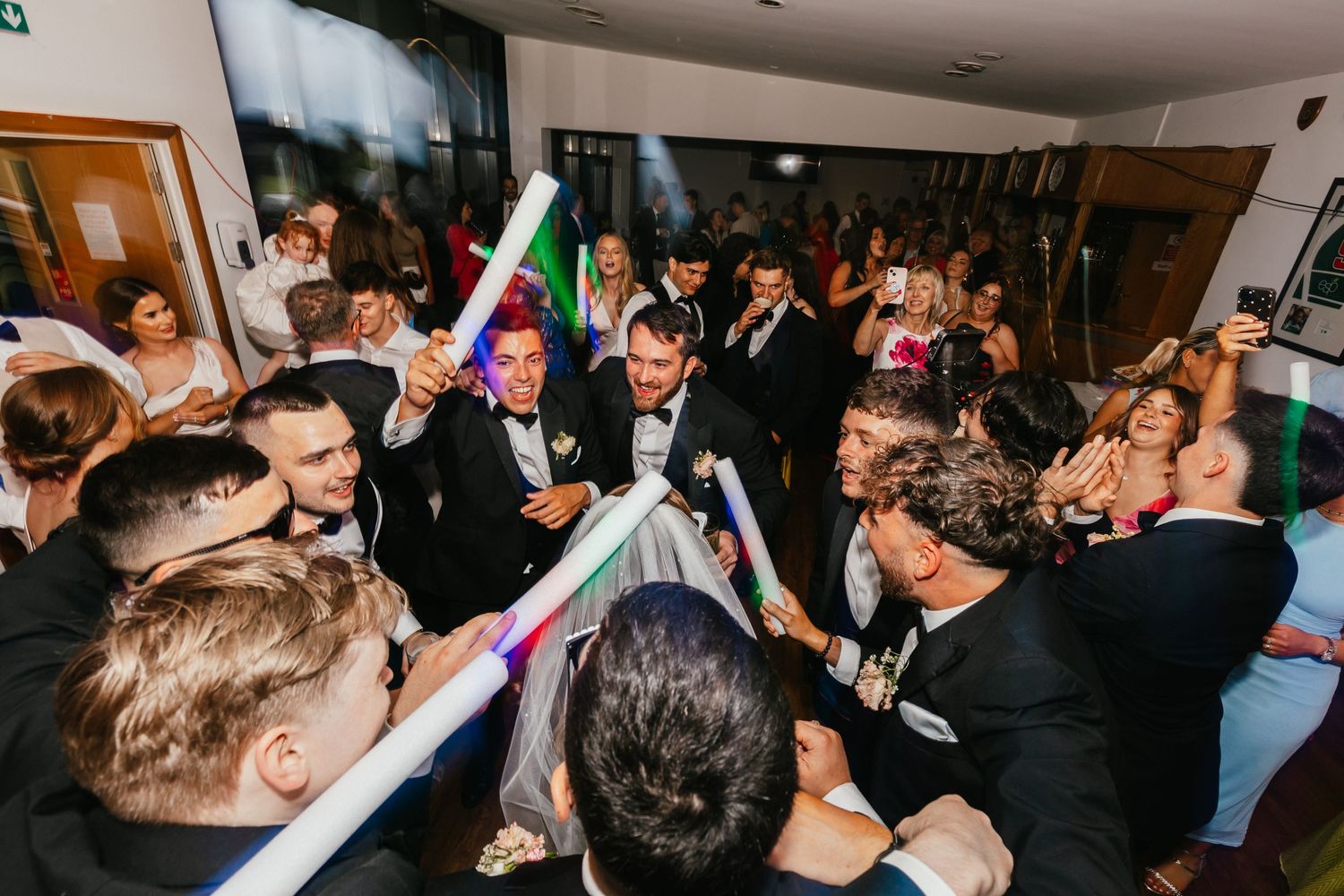Wedding guests wave light-up sticks while celebrating on a crowded dance floor.