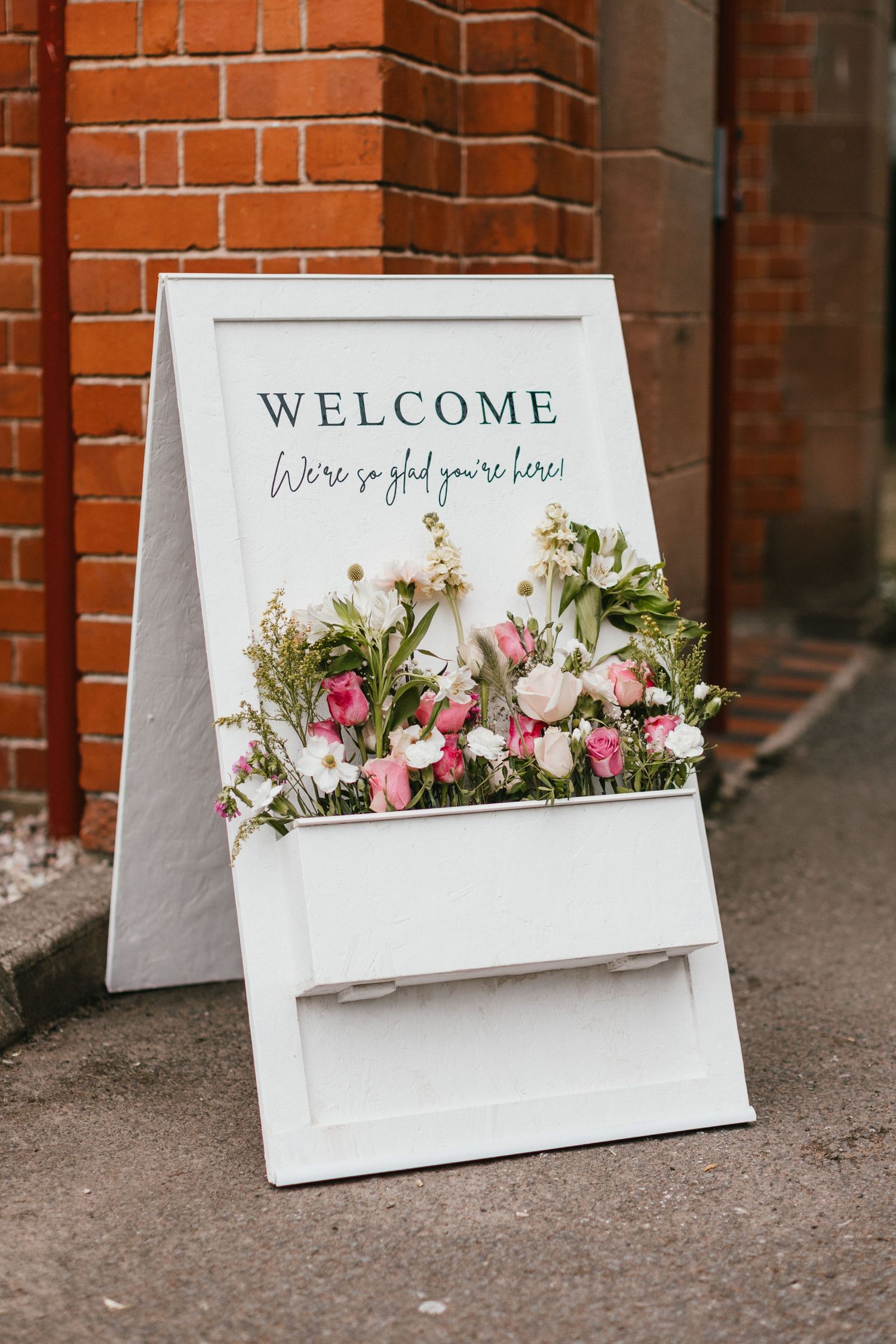 White welcome sign decorated with pink and white flowers stands against brick church wall.