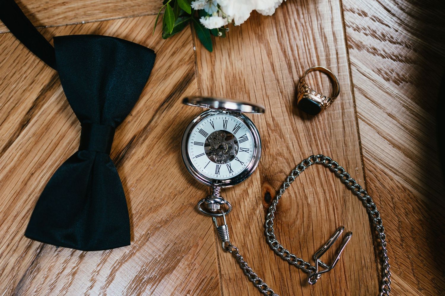 Black bow tie, vintage pocket watch, and ring arranged on wooden surface.