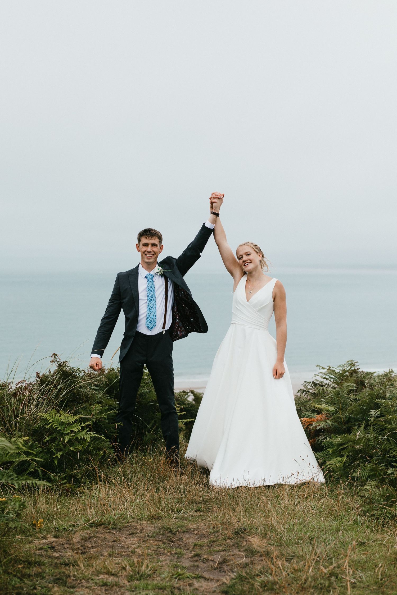 Celebratory wedding portrait of couple raising joined hands triumphantly on coastal clifftop.