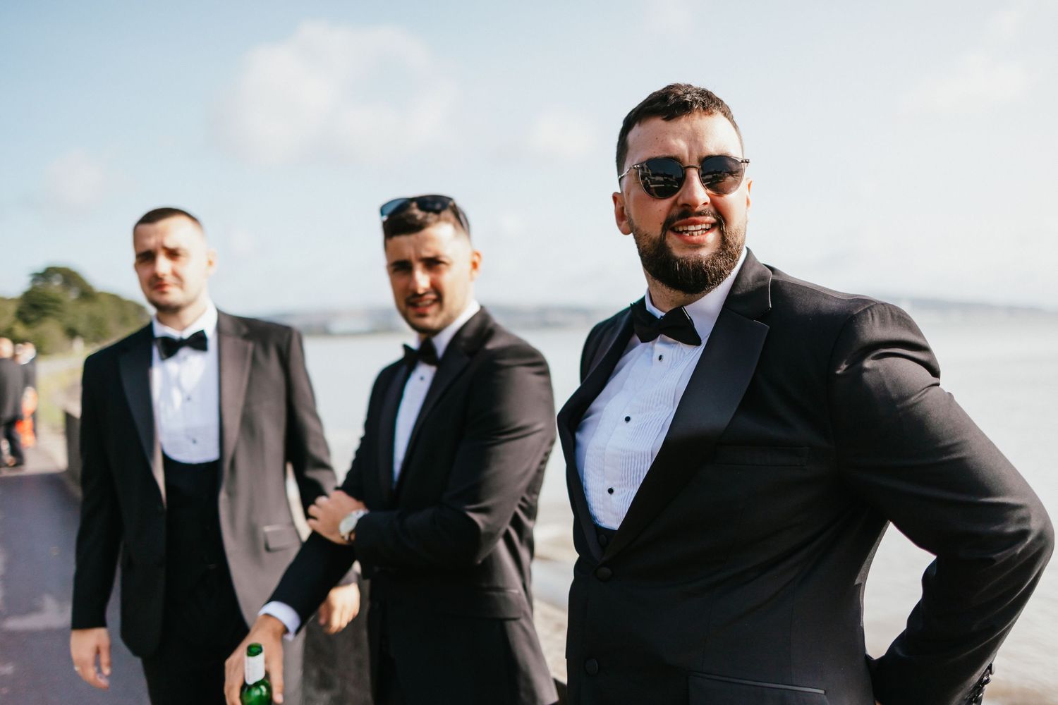 Groomsmen in black tuxedos and sunglasses pose together on a sunny day by the water.
