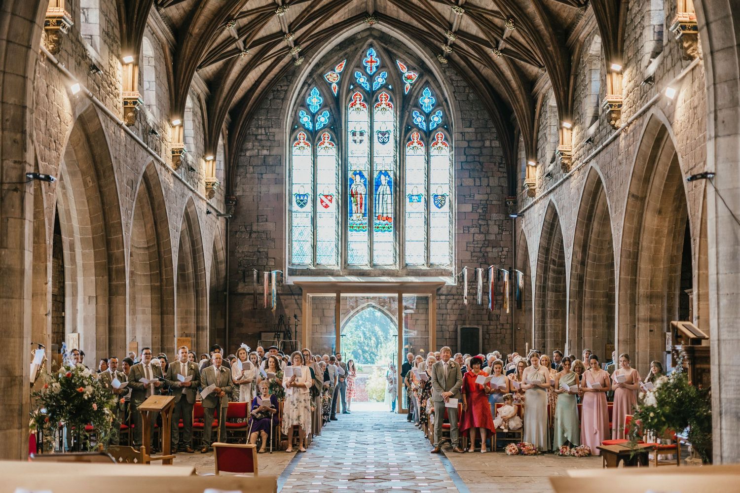 Beautiful church wedding ceremony in St Asaph Cathedral in North Wales with stained glass windows and stone arches.