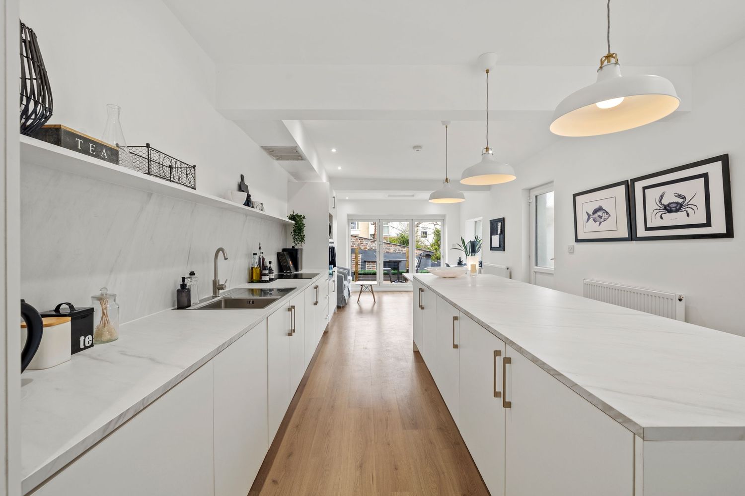 Long galley kitchen with white cabinets, pendant lights and wooden flooring extending to bright living space.