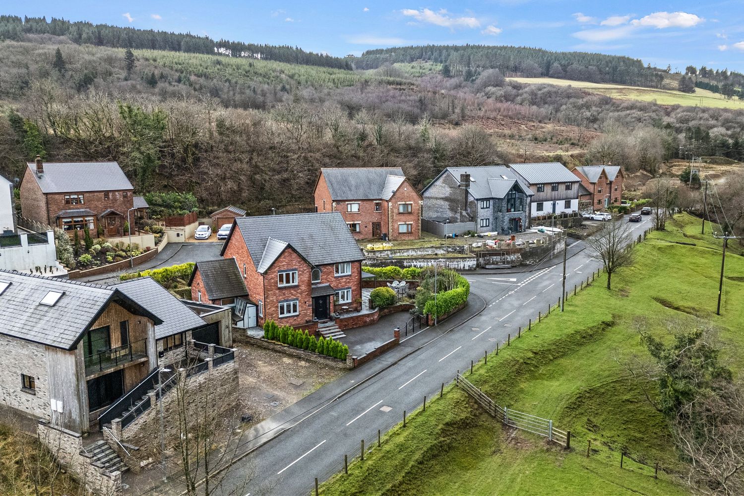 Aerial view of modern residential houses along a country road nestled in rolling hills with trees and greenery.