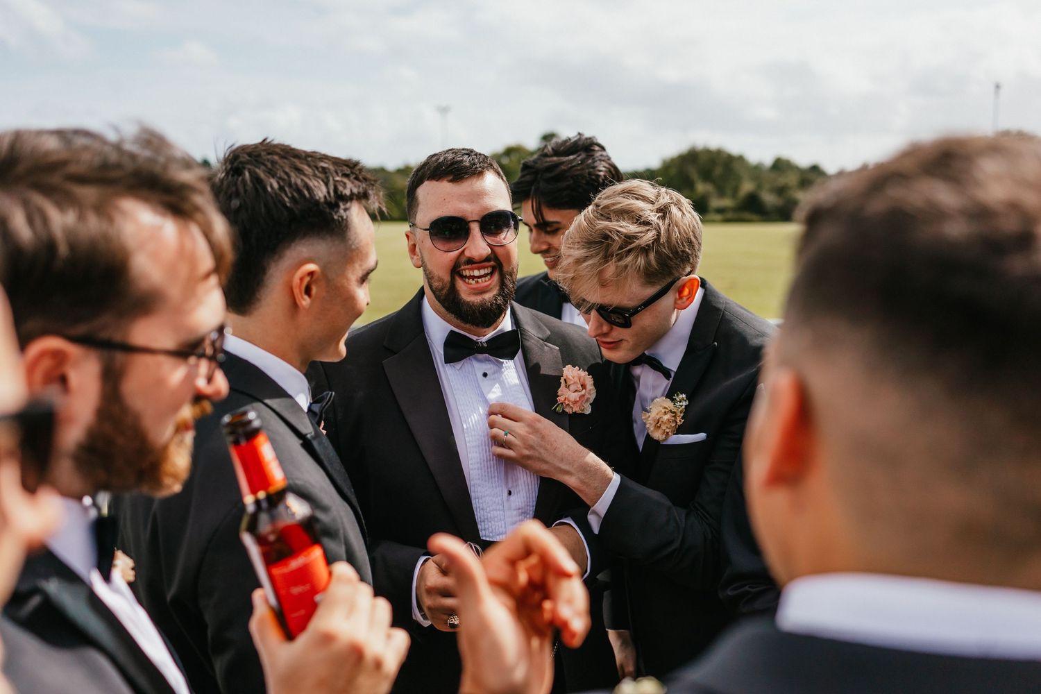 Groomsmen in formal attire gathered outdoors sharing drinks and conversation.