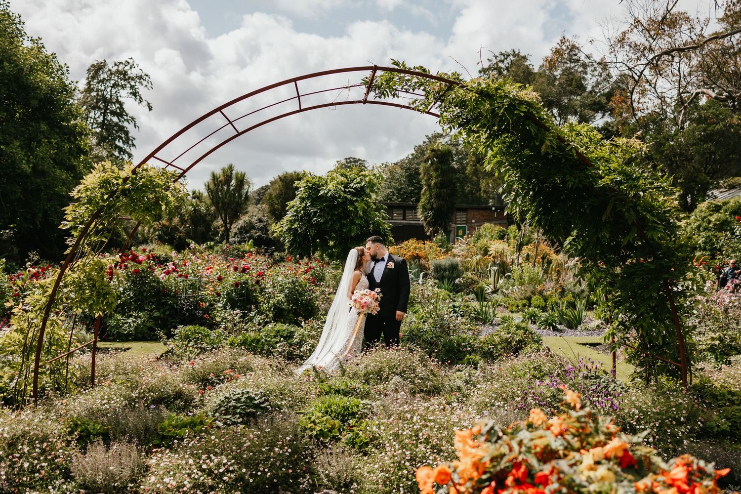 Wedding photograph under a rustic garden arch surrounded by blooming flowers.