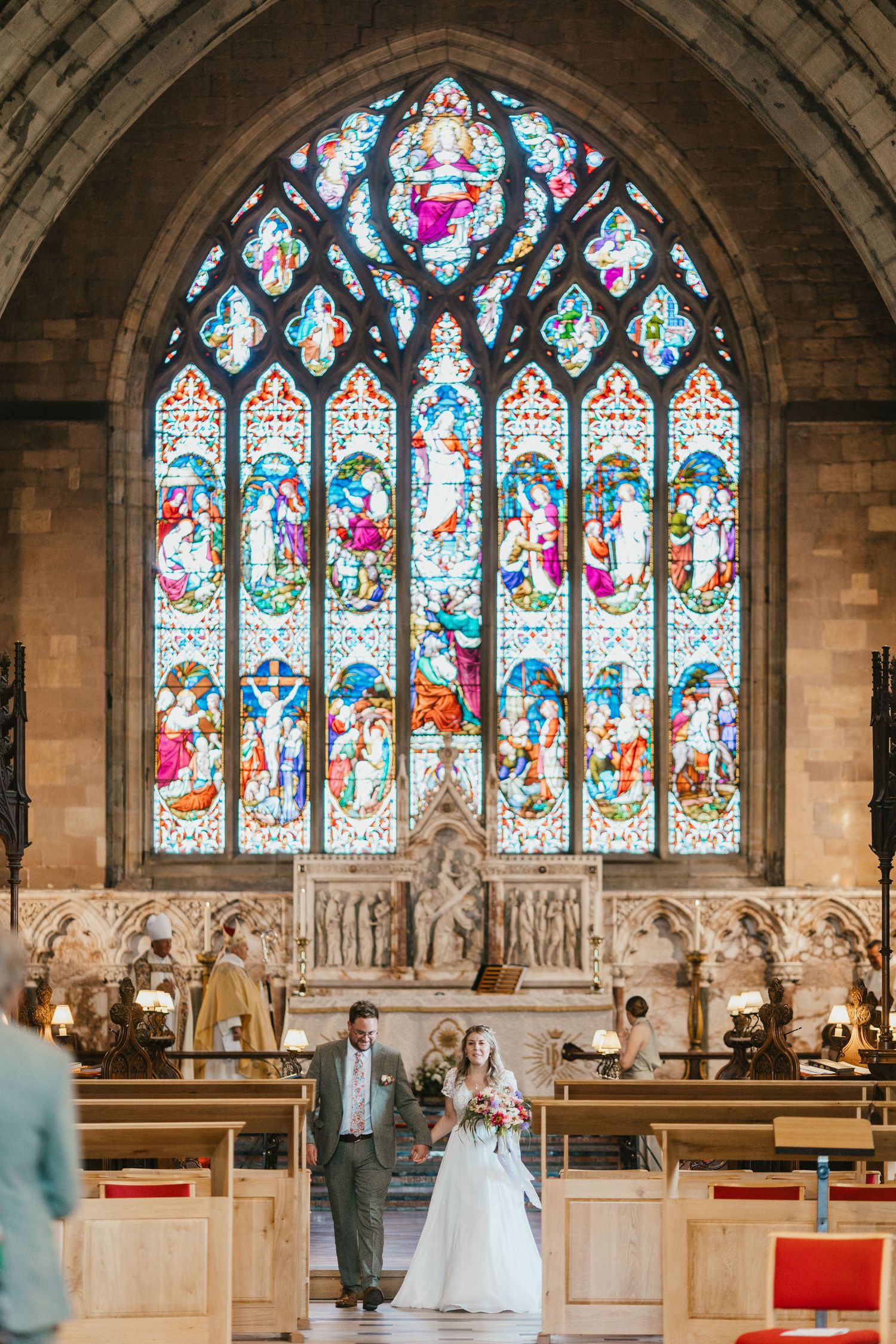 A wedding couple stands beneath a stunning stained glass church window with intricate religious artwork. St Asaph Cathedral.