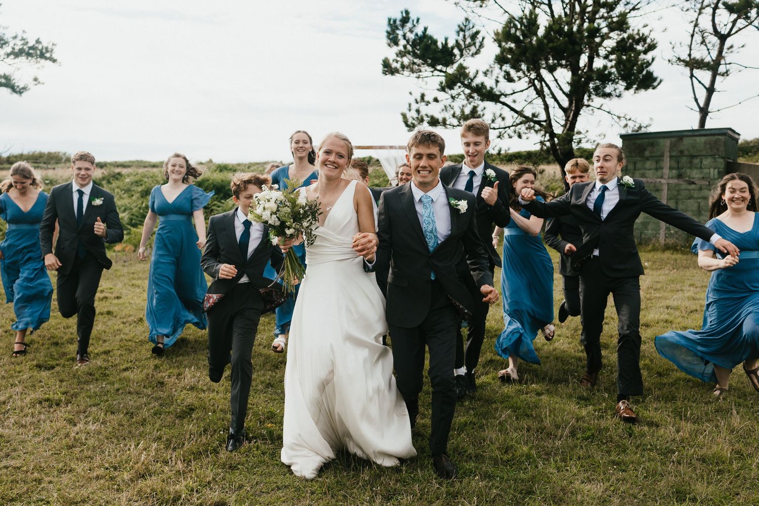 Joyful wedding party running and laughing together across a grassy field during outdoor celebration.