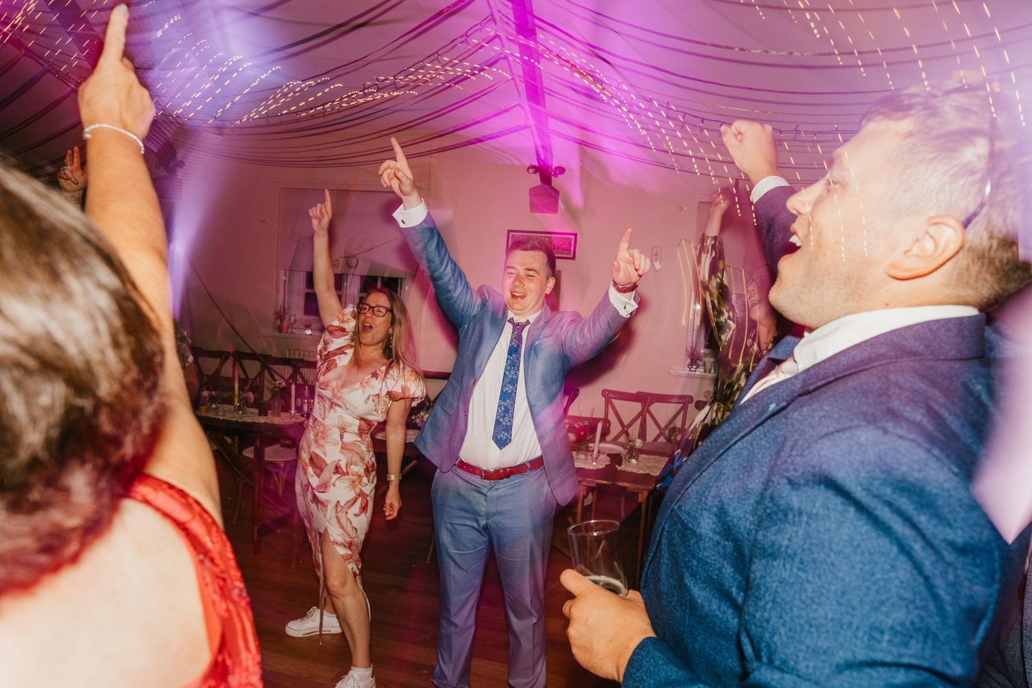 Guests celebrate with raised arms under purple lighting at a wedding reception.